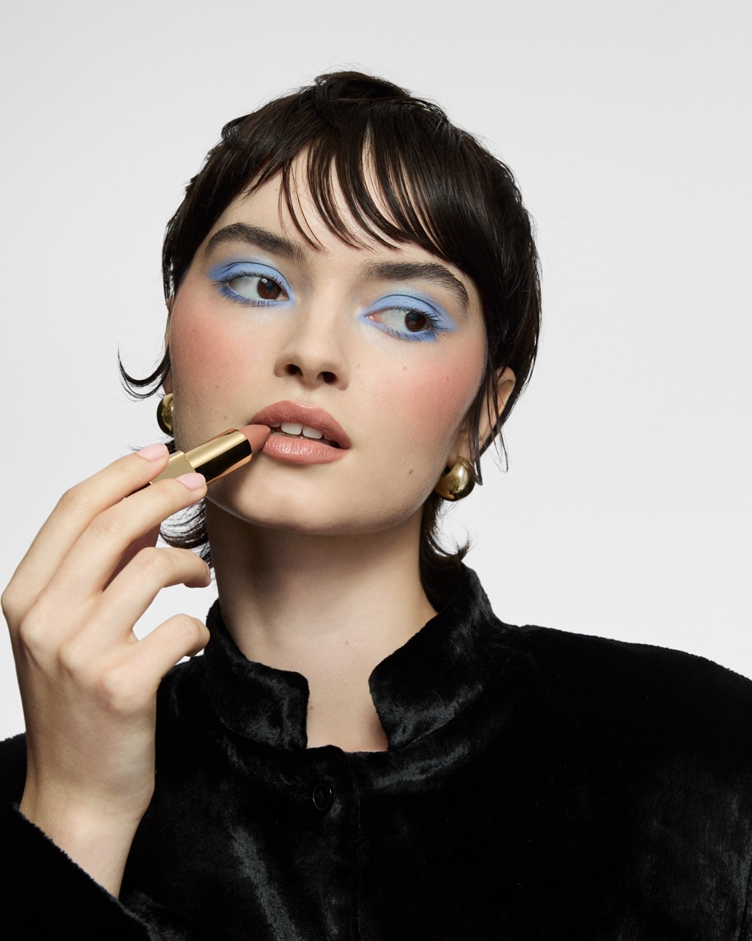 Eine Frau mit blauem Lidschatten und rosa Rouge trägt einen nudefarbenen Lippenstift aus einer goldenen Hülse auf.