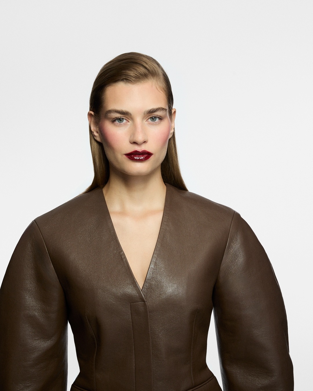 Eine Frau mit hellbraunem Haar, burgunderrotem Lippenstift und braunem Lederkleid posiert vor einem weißen Hintergrund.