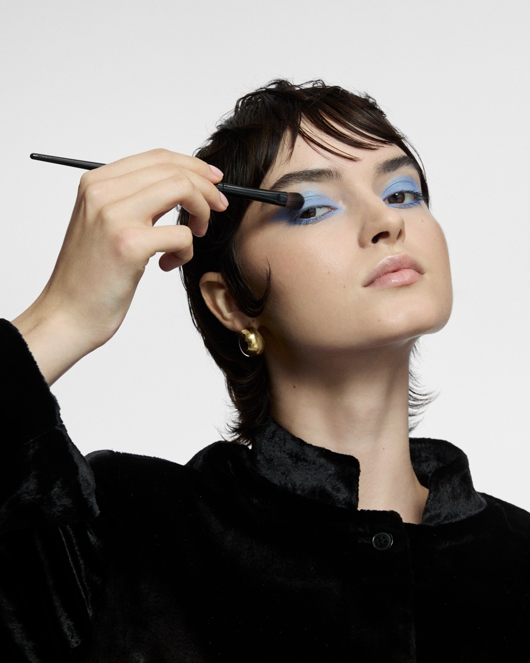 Eine Frau mit kurzem, dunklem Haar trägt mit einem Pinsel hellblauen Lidschatten auf ihr Augenlid auf, während sie einen schwarzen Samtmantel und goldene Ohrringe trägt.