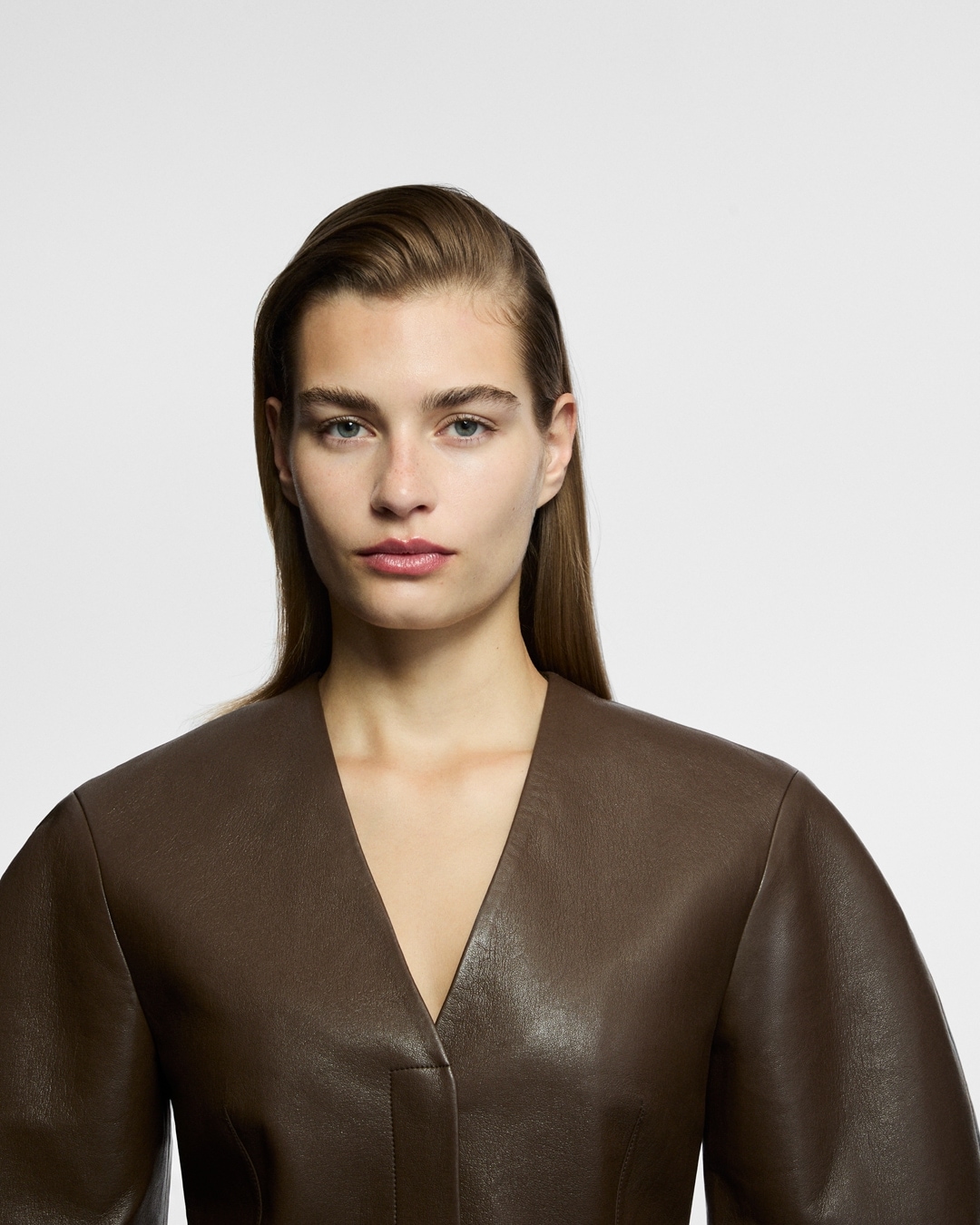 Nahaufnahme einer Frau mit glattem, braunem Haar und hellblauen Augen, die ein braunes Lederoberteil mit V-Ausschnitt trägt.