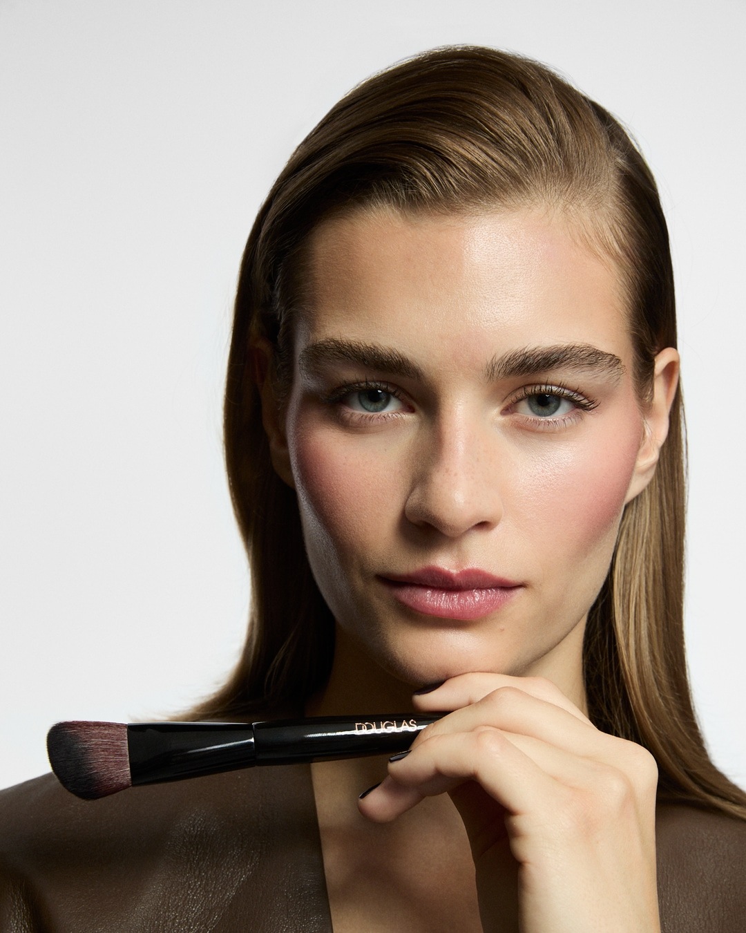 Eine Frau mit glatten, braunen Haaren und rosigem Rouge hält einen schwarzen Douglas-Make-up-Pinsel unter ihrem Kinn.