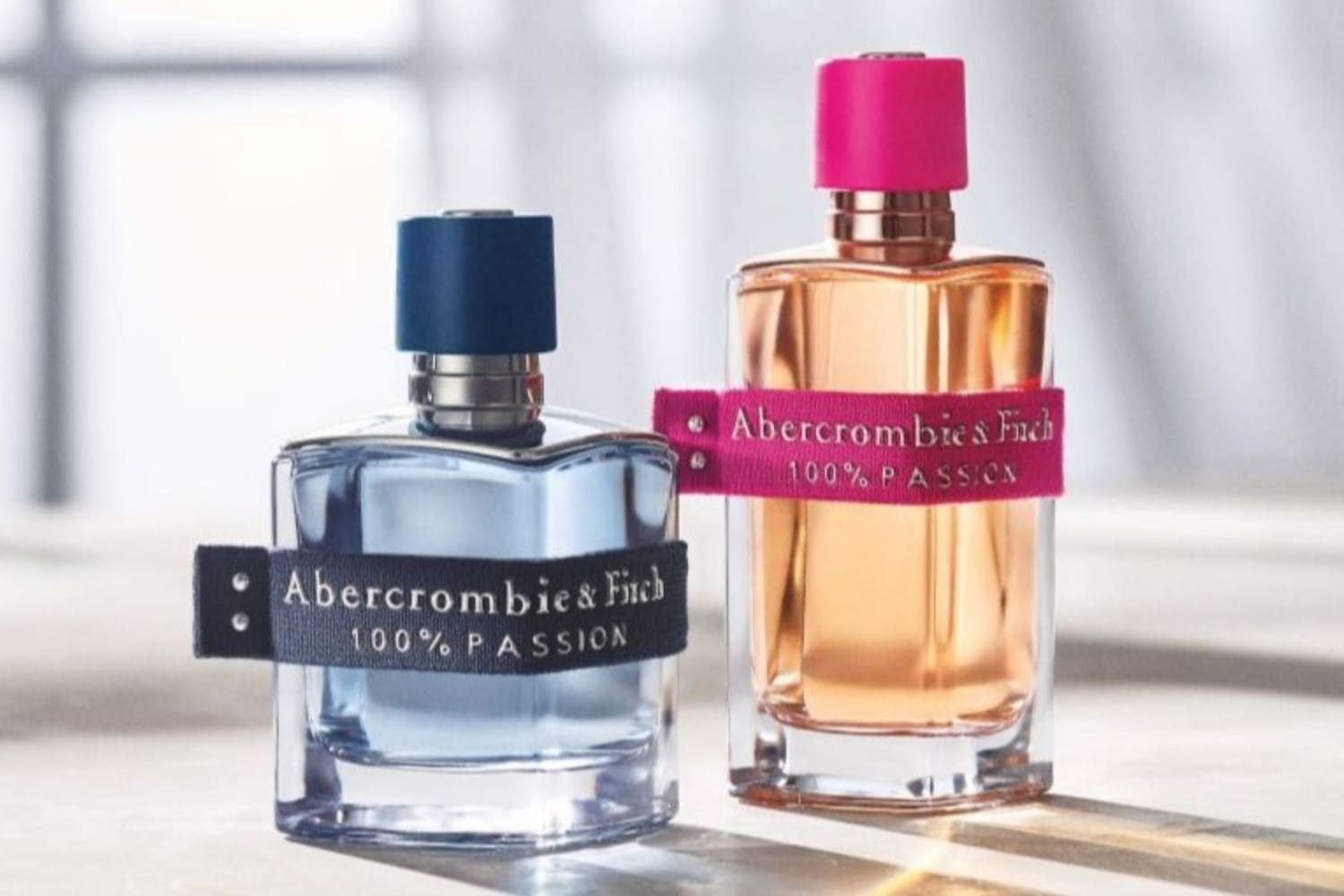Twee flessen Abercrombie & Fitch 100% PASSION parfum, één met blauwe dop en blauwe vloeistof, de andere met roze dop en perzikkleurige vloeistof.