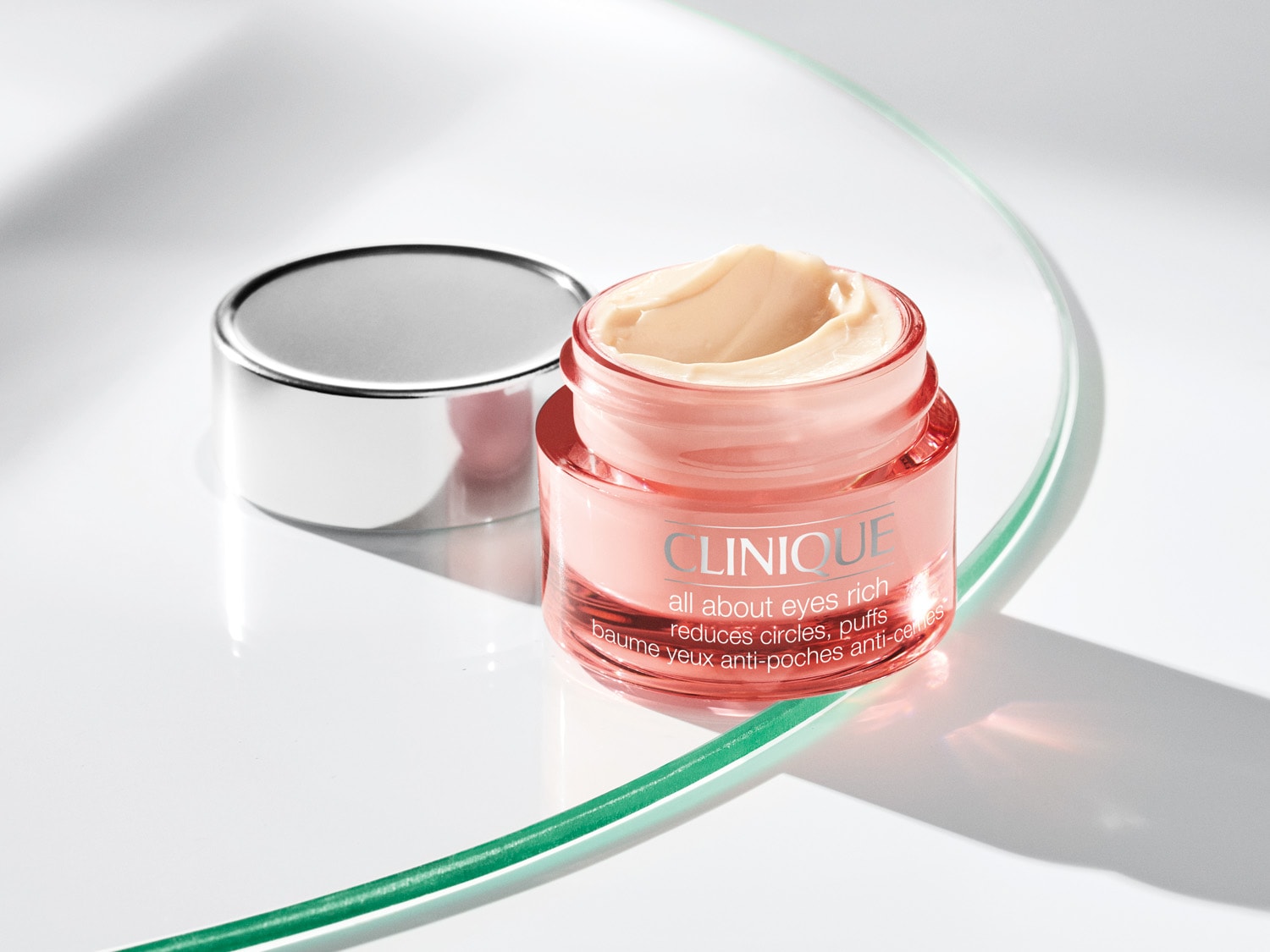 Die Clinique All About Eyes Rich Creme in einem durchsichtigen, rosafarbenen Tiegel mit silbernem Deckel ist auf Glas platziert.