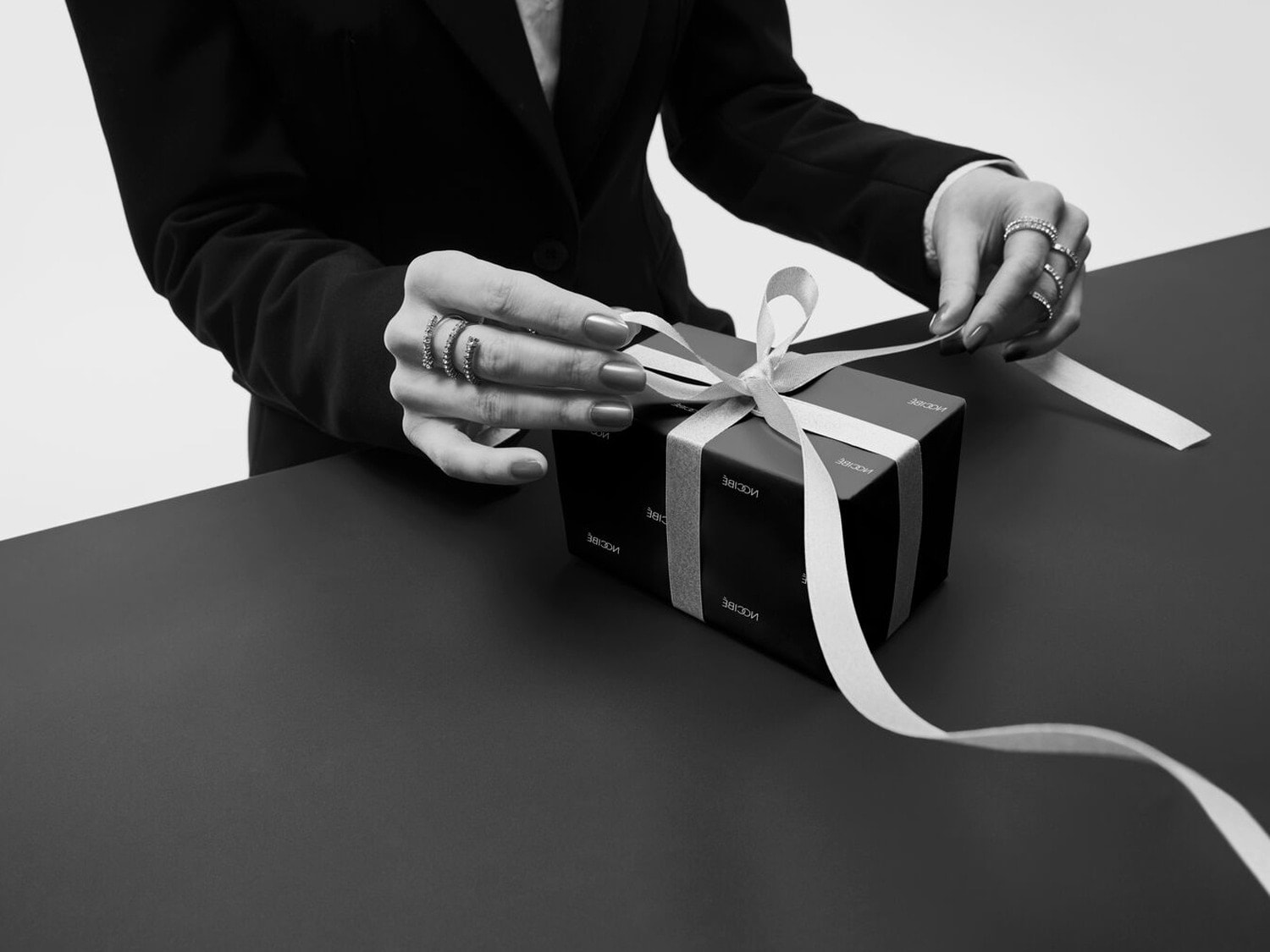 Gros plan en noir et blanc d'une personne en costume ouvrant un cadeau emballé dans du papier noir et un ruban blanc, mettant en valeur l'anticipation d'un moment spécial.