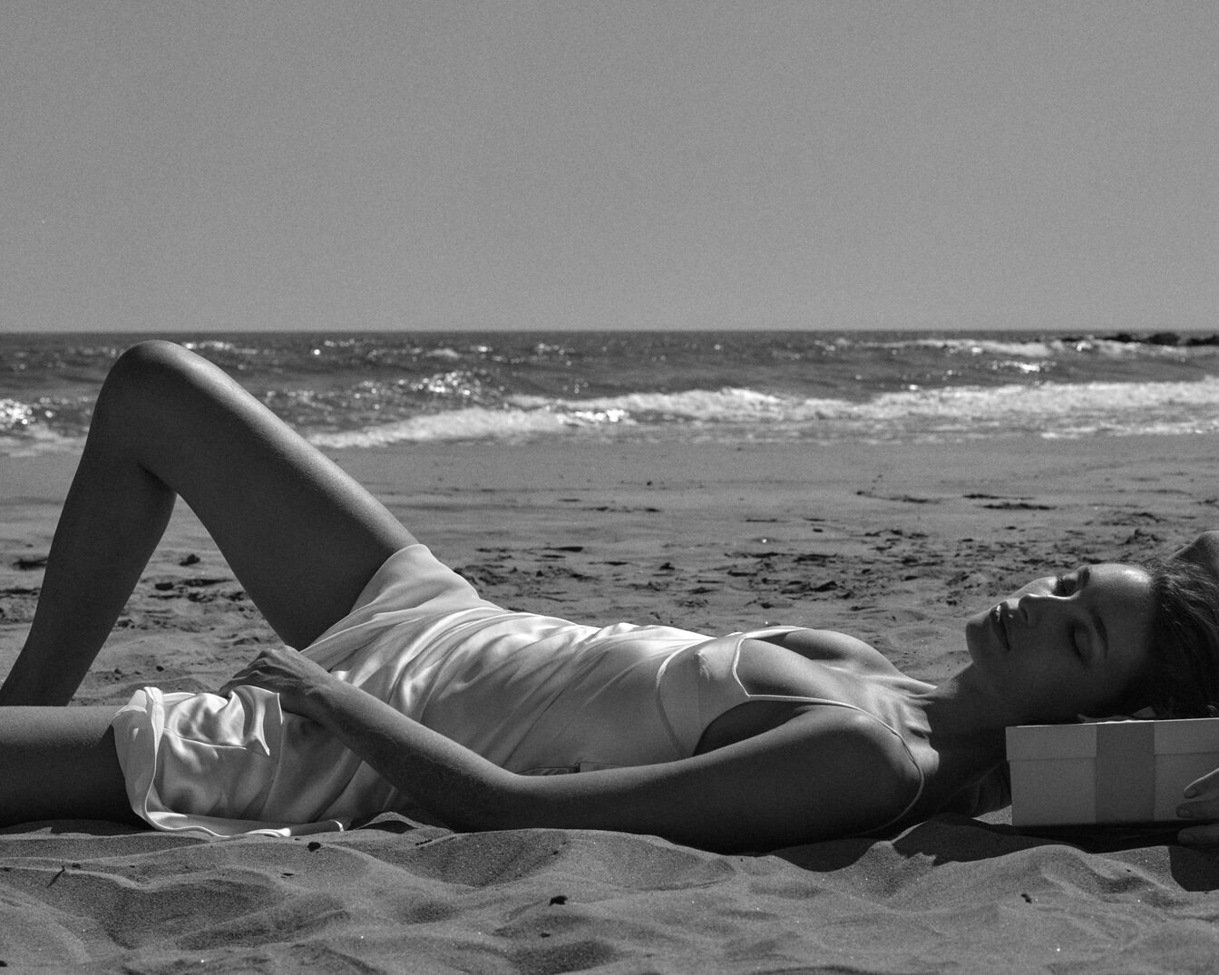 Une femme en maillot de bain clair se détend sur une plage de sable, les yeux fermés, avec une boîte cadeau à côté d'elle et la mer en arrière-plan.