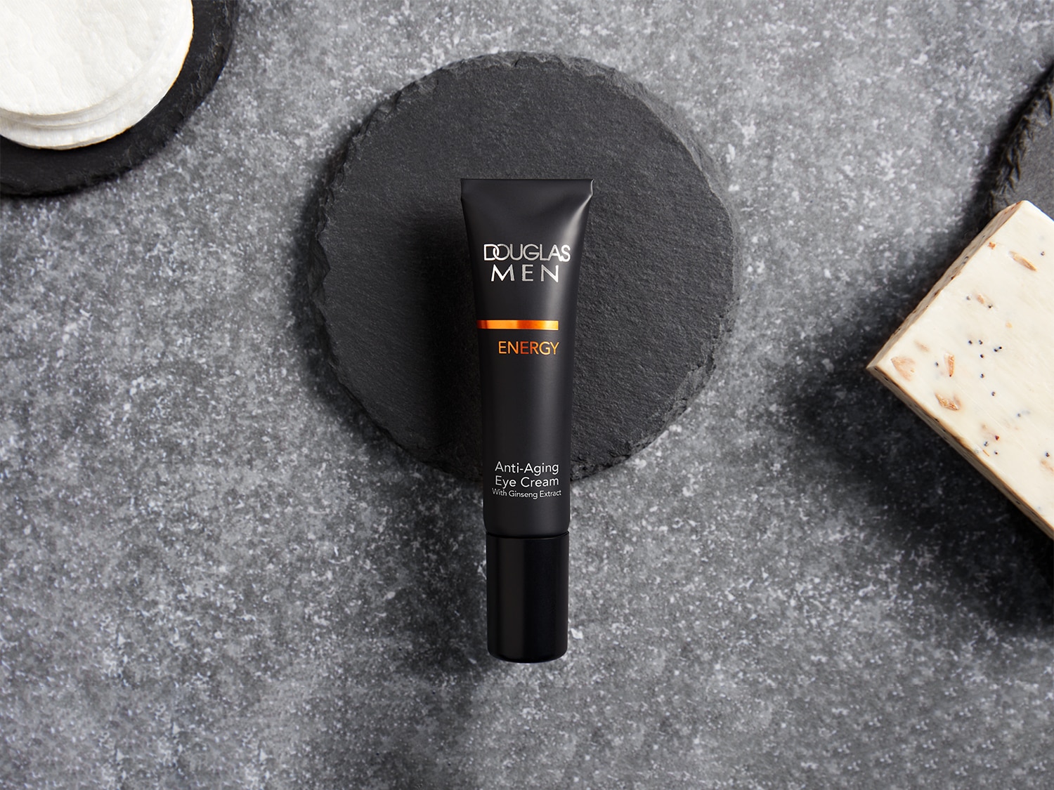 Voici un tube noir de crème contour des yeux anti-âge Douglas Men Energy, enrichie à l'extrait de ginseng, posé sur un fond gris texturé.