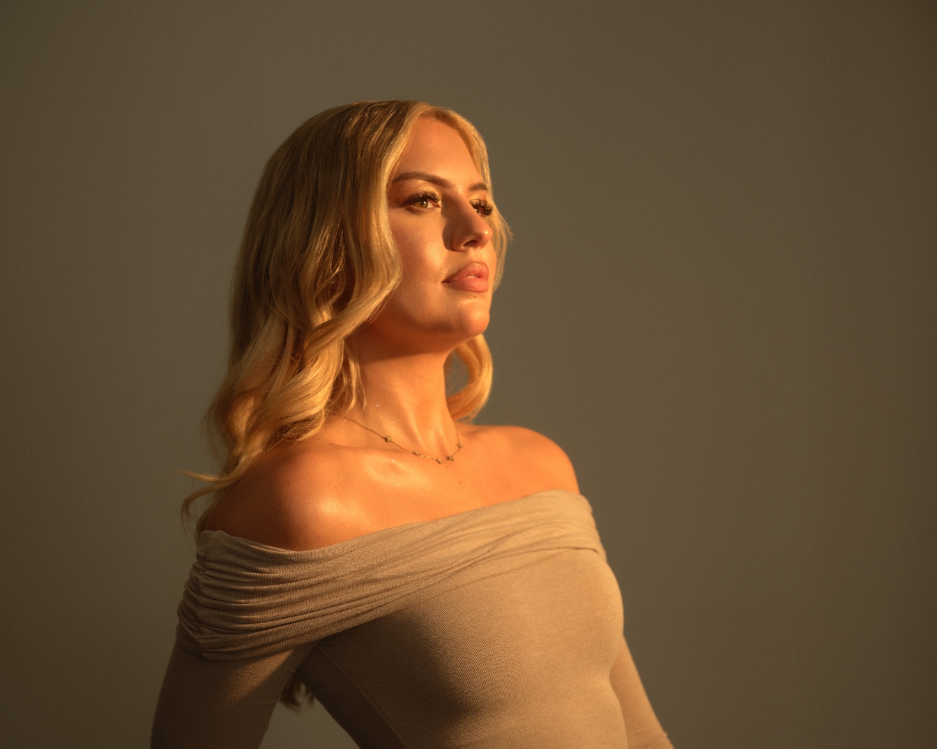 Eine blonde Frau mit gewelltem Haar und einem schulterfreien Oberteil blickt in einem warmen Licht nach oben.