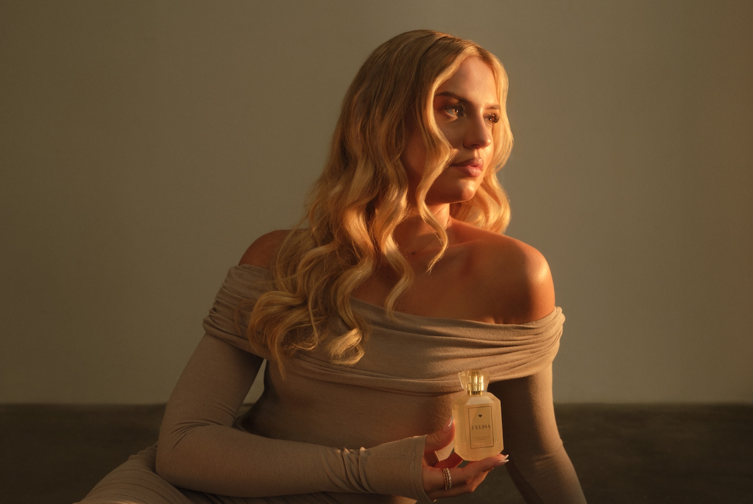 Blonde Frau im beigen Kleid, die eine Flasche J'ELISA-Parfüm mit goldfarbenem Verschluss hält.