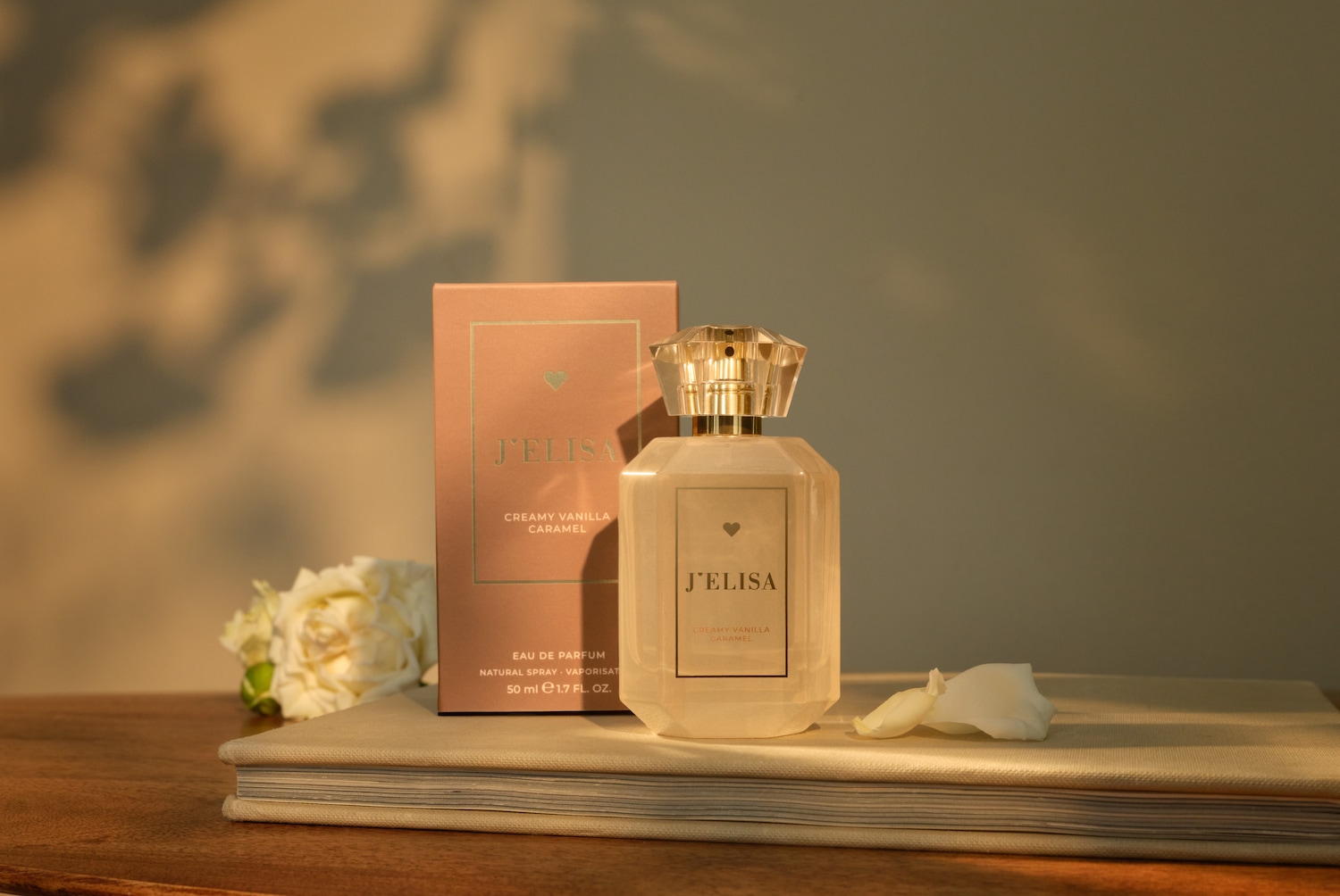 J'ELISA Creamy Vanilla Caramel Eau de Parfum in einer klaren Flasche mit facettiertem Verschluss und rosa Verpackung, die auf einem Buch mit weißen Rosen liegt.