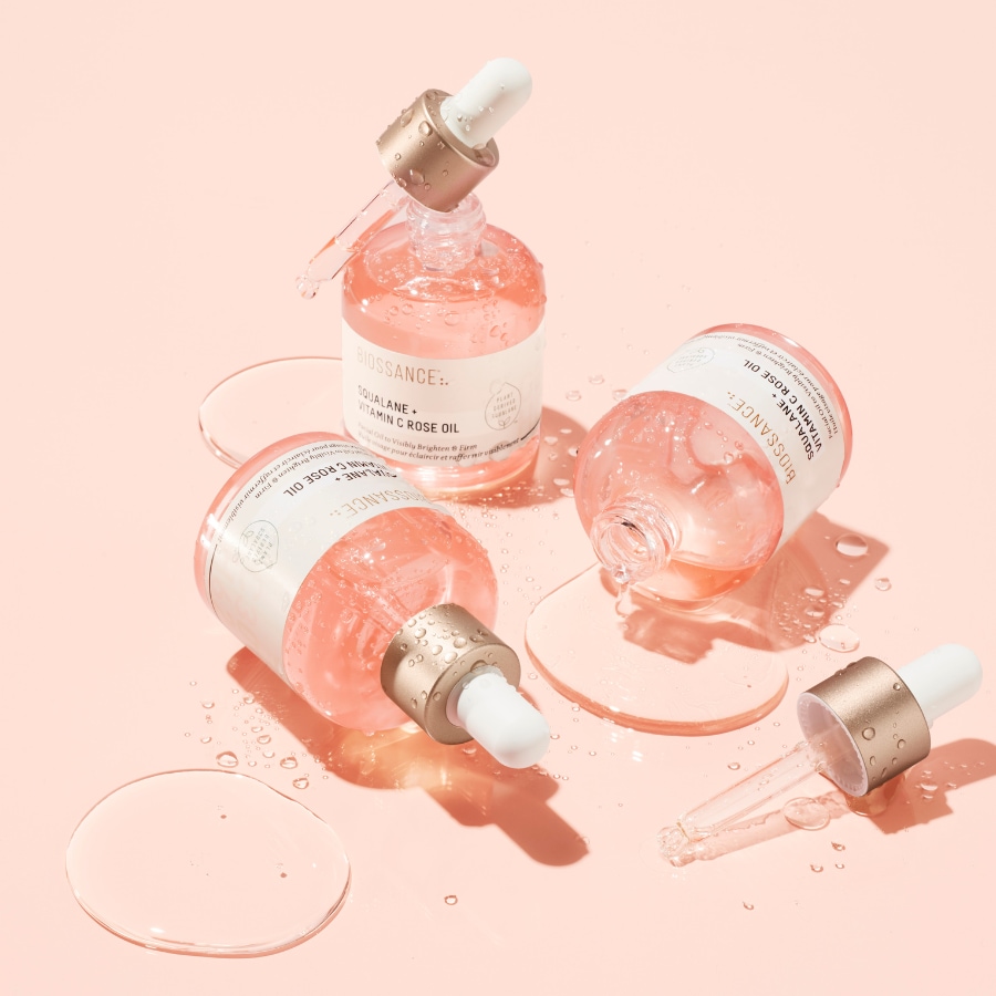 Nahaufnahme von drei Flaschen Biossance Squalane + Vitamin C Rose Oil mit Pipetten auf einem hellrosa Hintergrund, die die feuchtigkeitsspendende und aufhellende Wirkung des Produkts hervorheben.