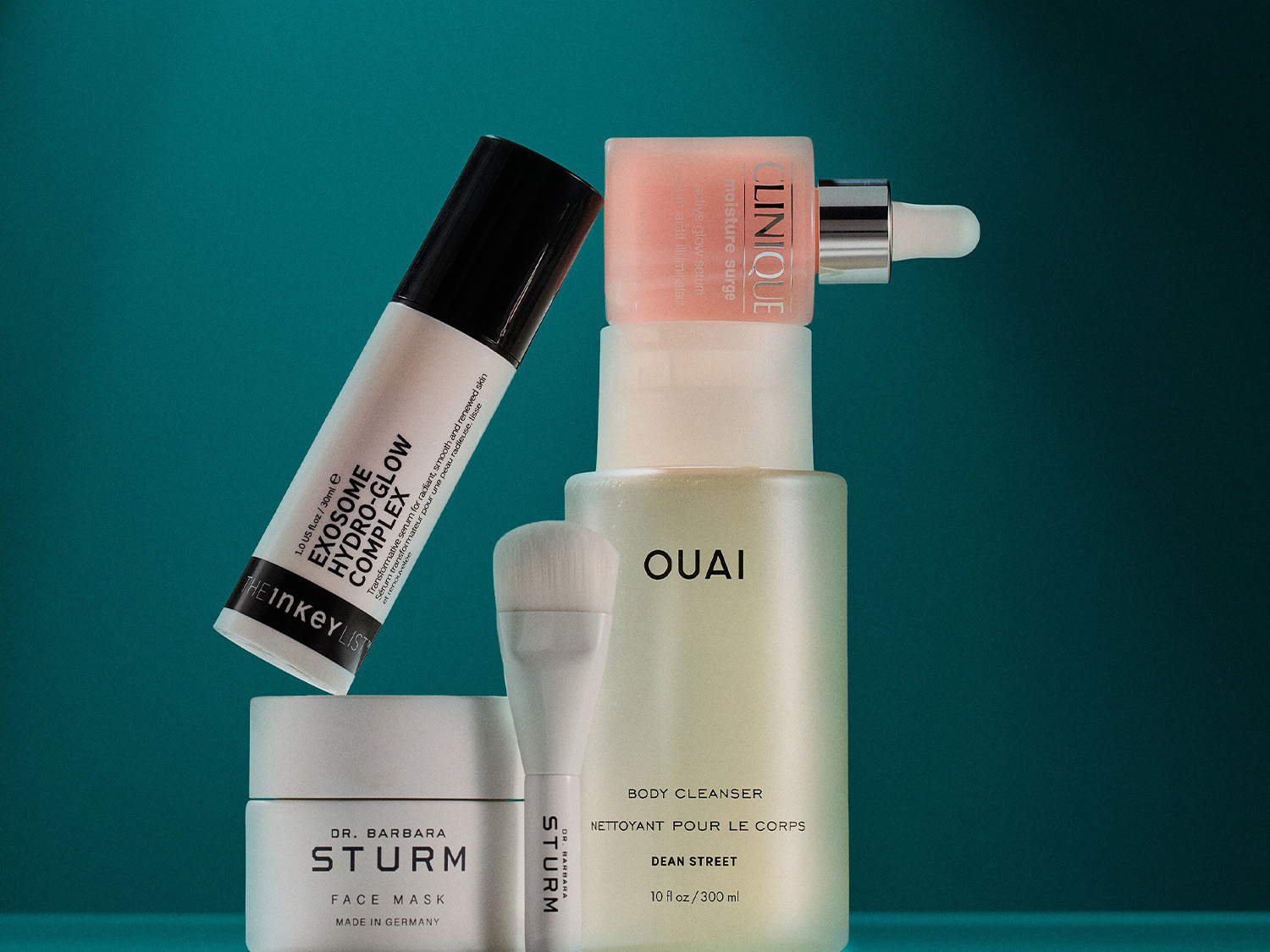 Verschiedene Hautpflegeprodukte, darunter The Inkey List Exosome Hydro-Glow Complex, Clinique Moisture Surge und OUAI Body Cleanser, die auf einem türkisfarbenen Hintergrund angeordnet sind.