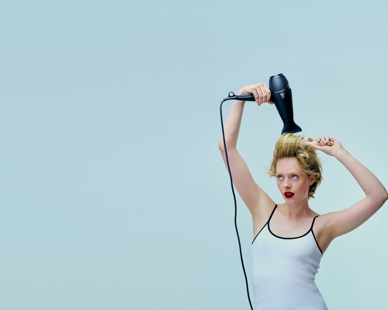 Eine Frau mit roten Lippen und blonden Haaren föhnt ihre Haare mit einem schwarzen Föhn vor einem hellblauen Hintergrund.