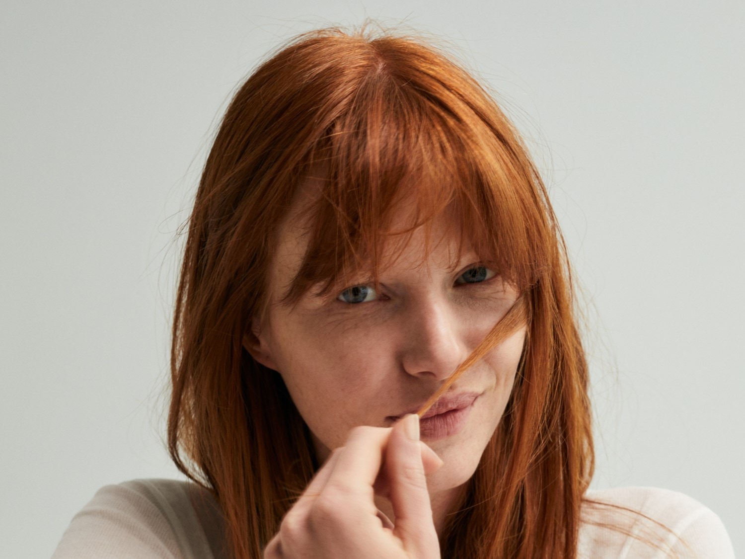 Nahaufnahme einer Frau mit langen, roten Haaren, die verspielt eine Haarsträhne vor ihr Gesicht hält und dabei leicht lächelt.
