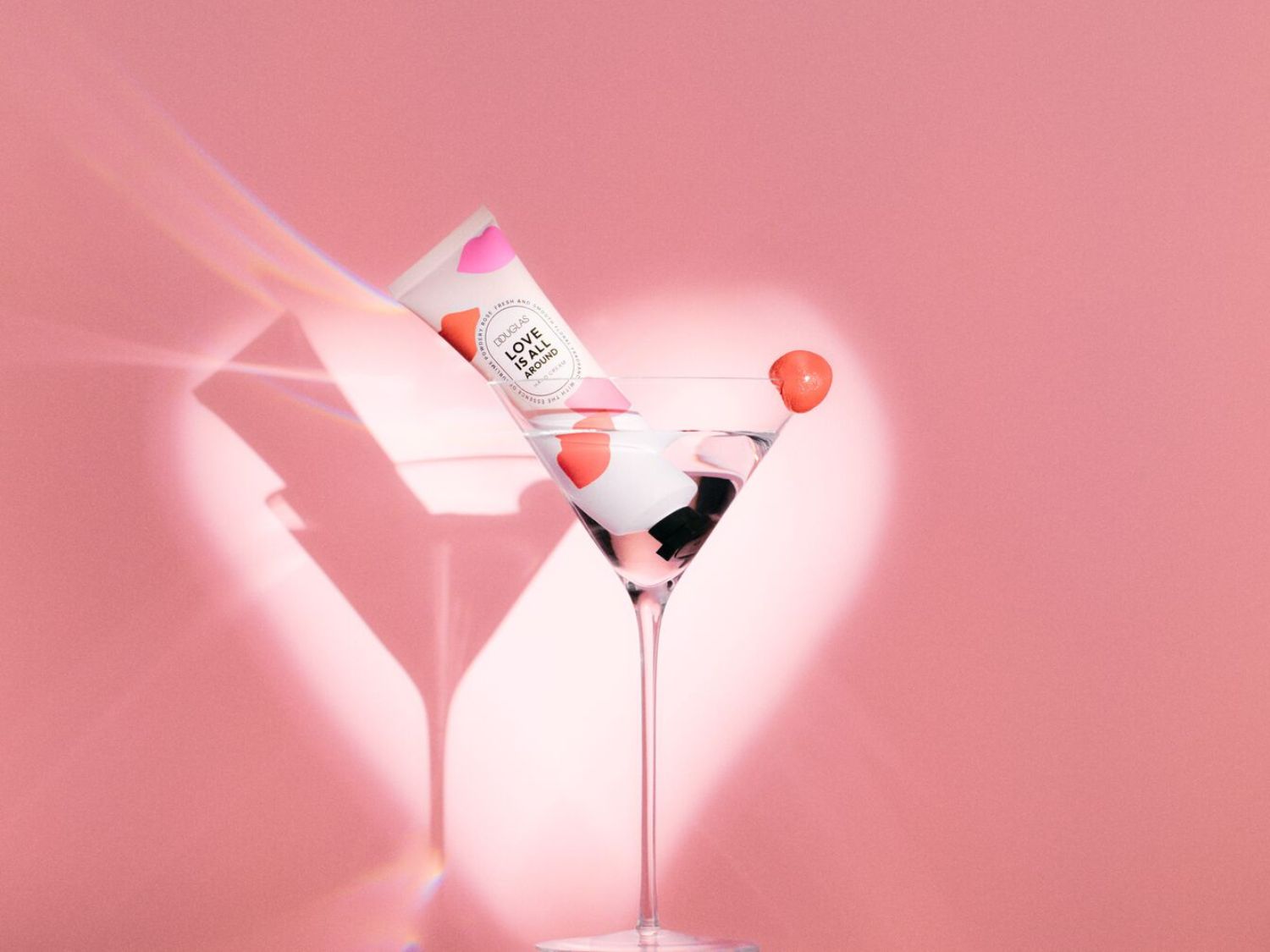 Le tube de crème pour les mains Douglas « Love is All Around » est présenté dans un verre à cocktail sur un fond rose, évoquant une ambiance festive et soignée.