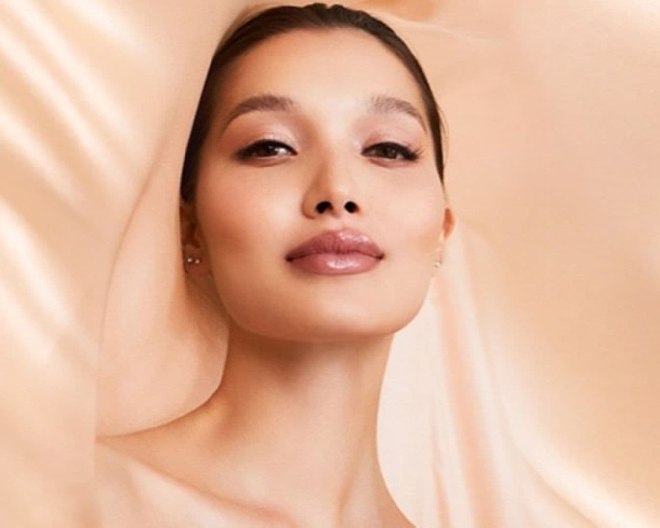 Nahaufnahme einer Frau mit glänzenden Lippen und zarter Haut vor einem weichen, pfirsichfarbenen Hintergrund.