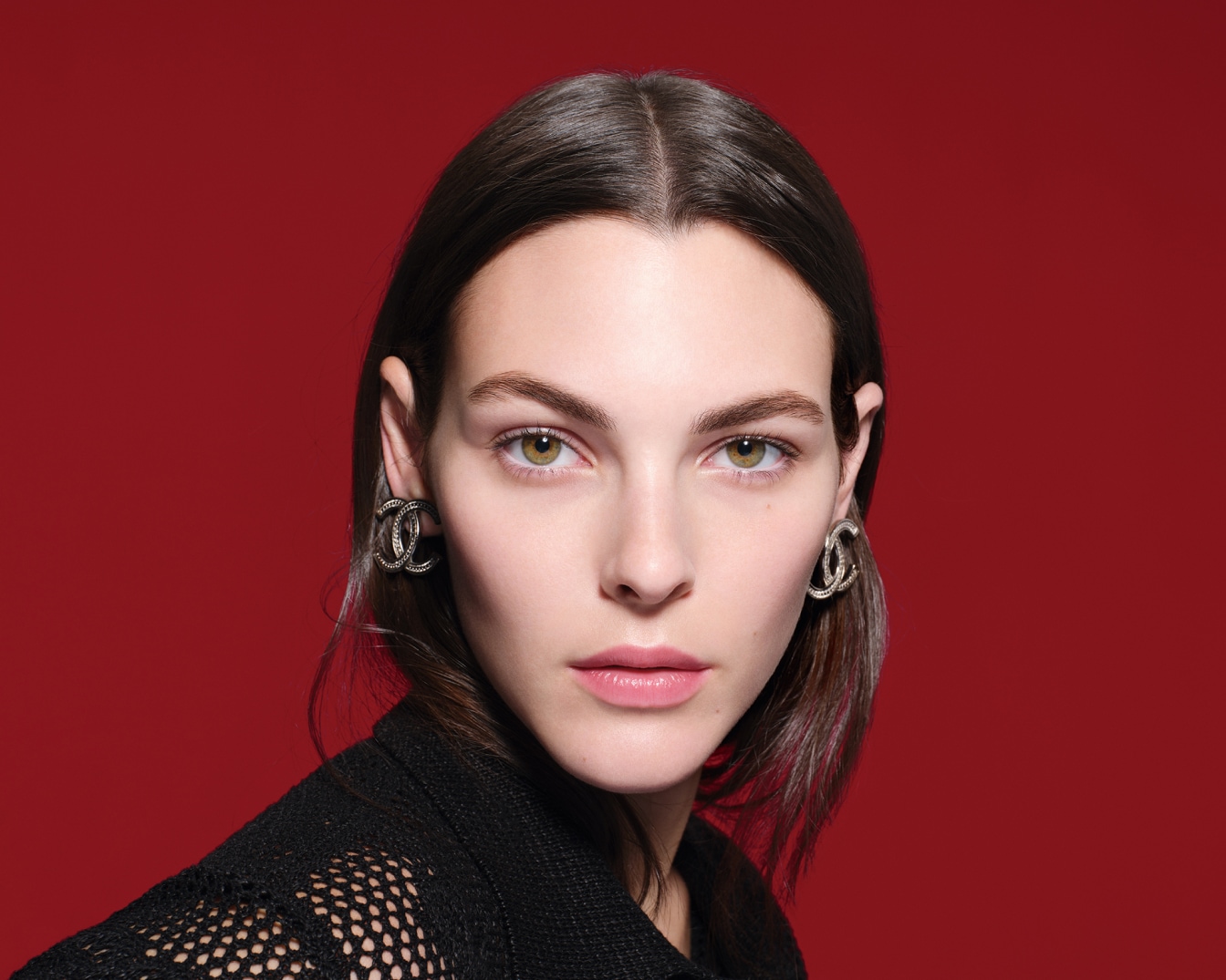 Close-up van een vrouw met bruin haar en groene ogen, met zilveren Chanel oorbellen tegen een rode achtergrond.