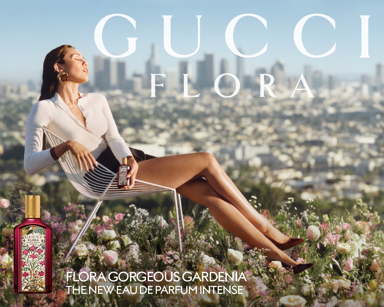 Une femme est assise sur une chaise au milieu des fleurs, tenant un flacon de parfum Gucci Flora Gorgeous Gardenia, avec le paysage urbain en arrière-plan.