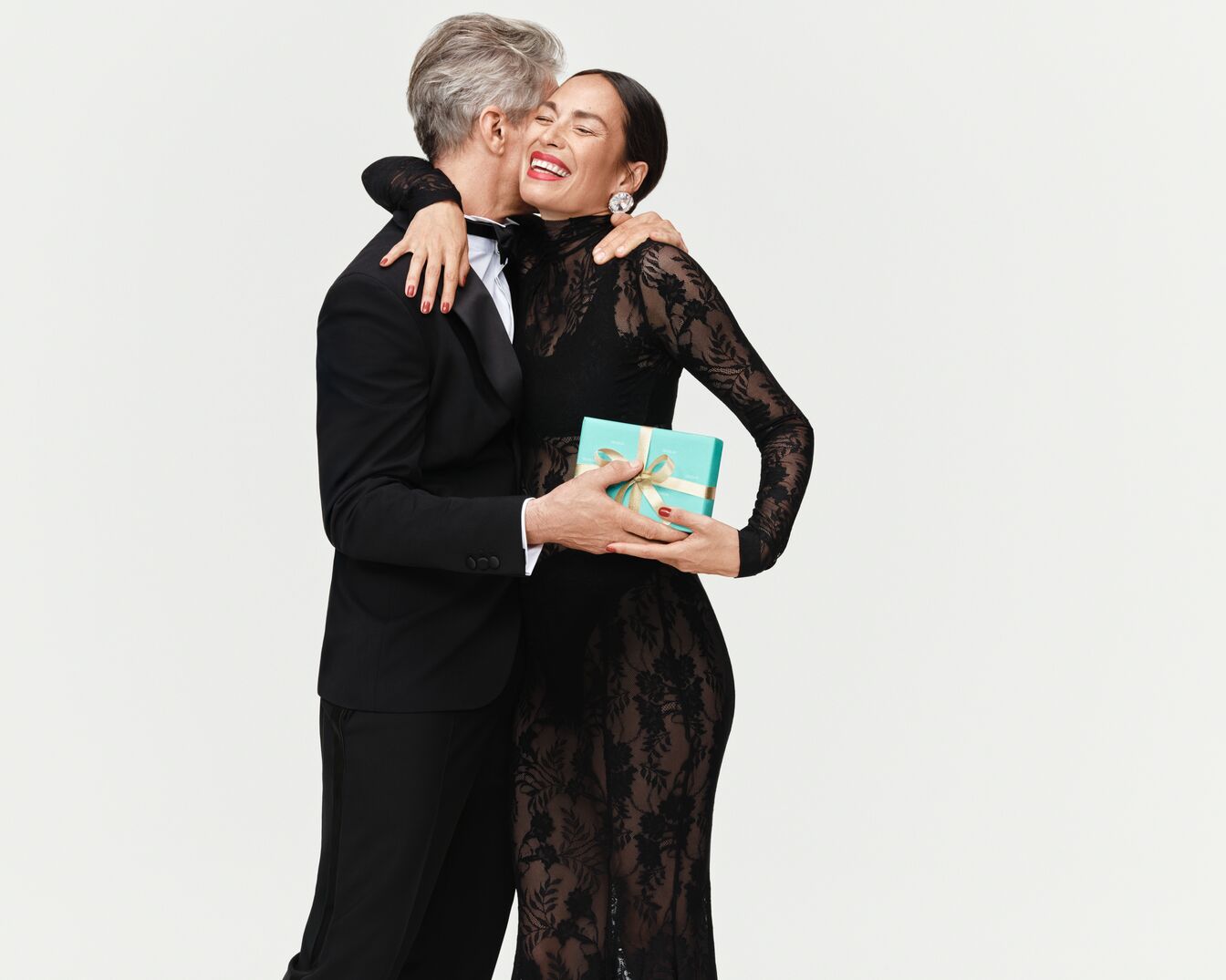 Ein Mann im Smoking umarmt eine Frau im schwarzen Spitzenkleid, die eine hellblaue Schachtel mit goldener Schleife hält.