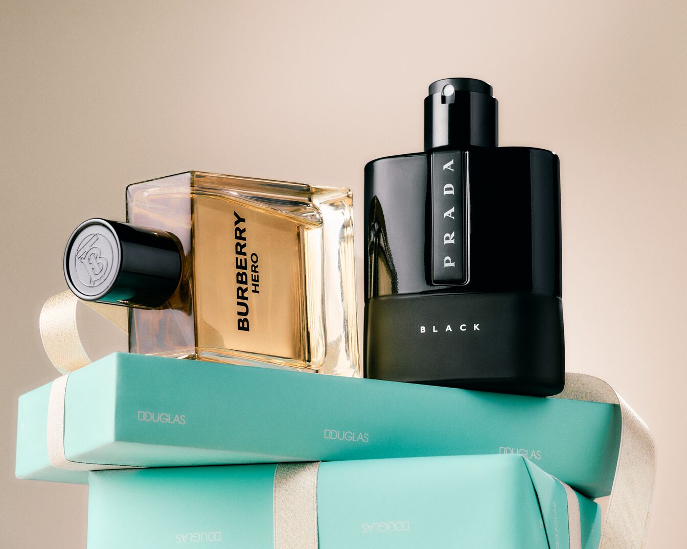 Ant mėlynų dovanų dėžučių su kaspinais pavaizduoti du kvepalų buteliukai: vienas auksinio skysčio stačiakampyje, pažymėtas „Burberry Hero“, o kitas – juodas, pažymėtas „Prada Black“.