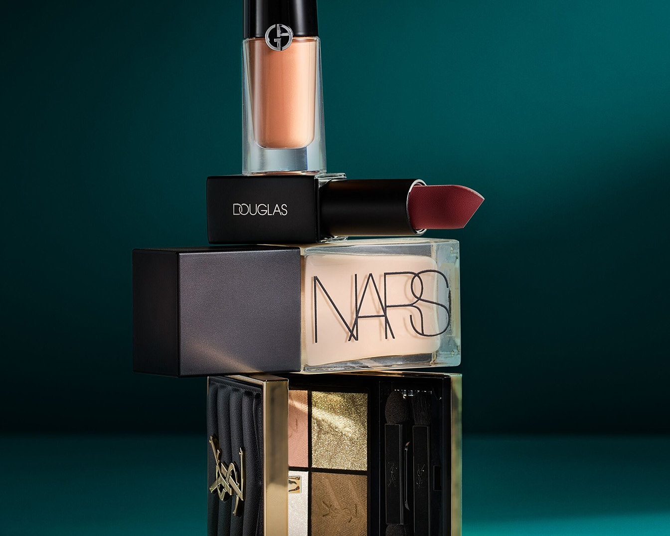 Auf einem grünen Hintergrund sind verschiedene Make-up-Produkte gestapelt, darunter ein goldfarbener Lipgloss von Giorgio Armani, ein Douglas Lippenstift, ein NARS-Produkt und eine goldene Lidschattenpalette von Yves Saint Laurent.