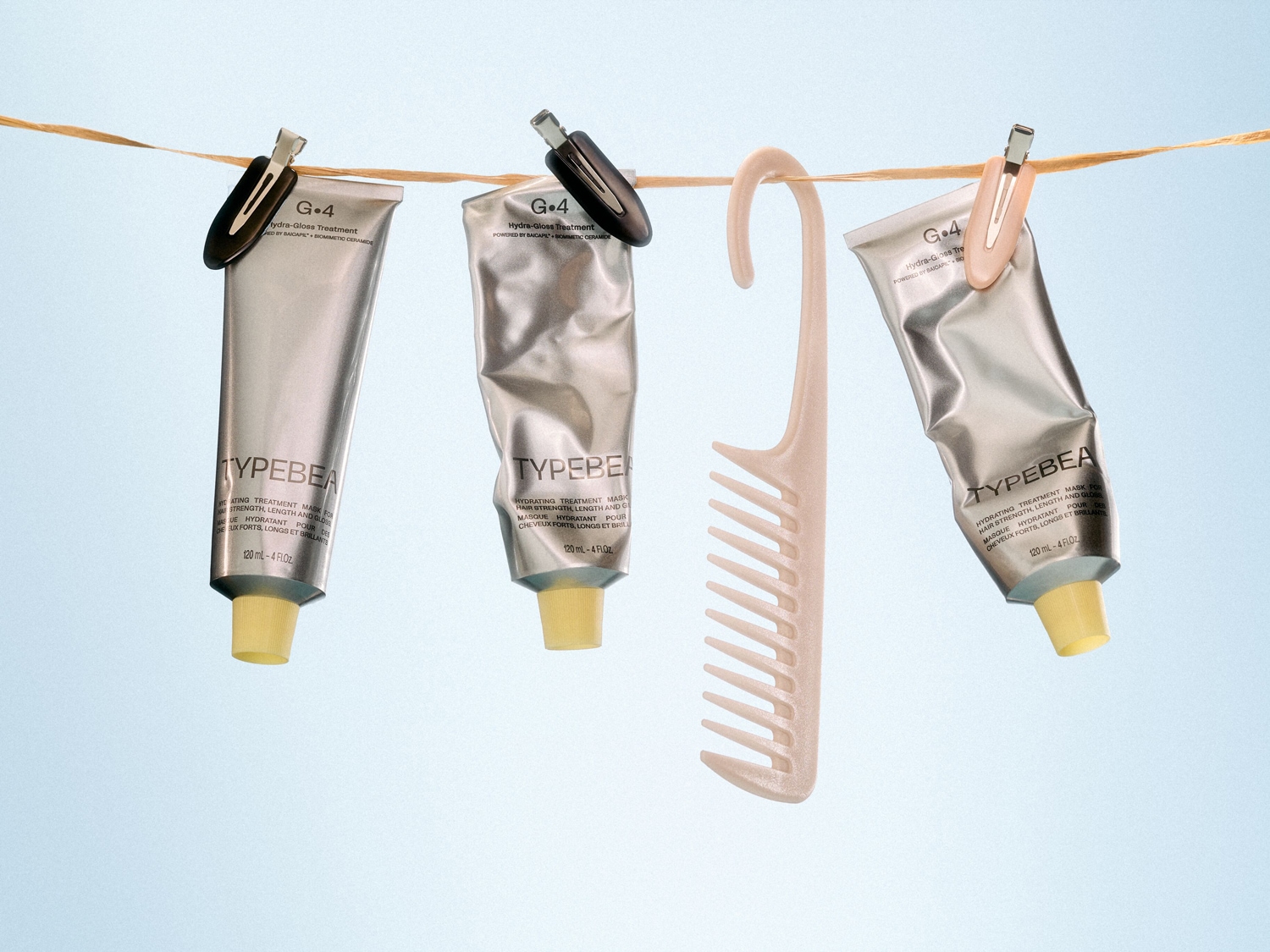 Trois tubes argentés de masque capillaire hydratant TYPEBEA G.4 sont suspendus à une corde à linge avec une brosse à cheveux beige.