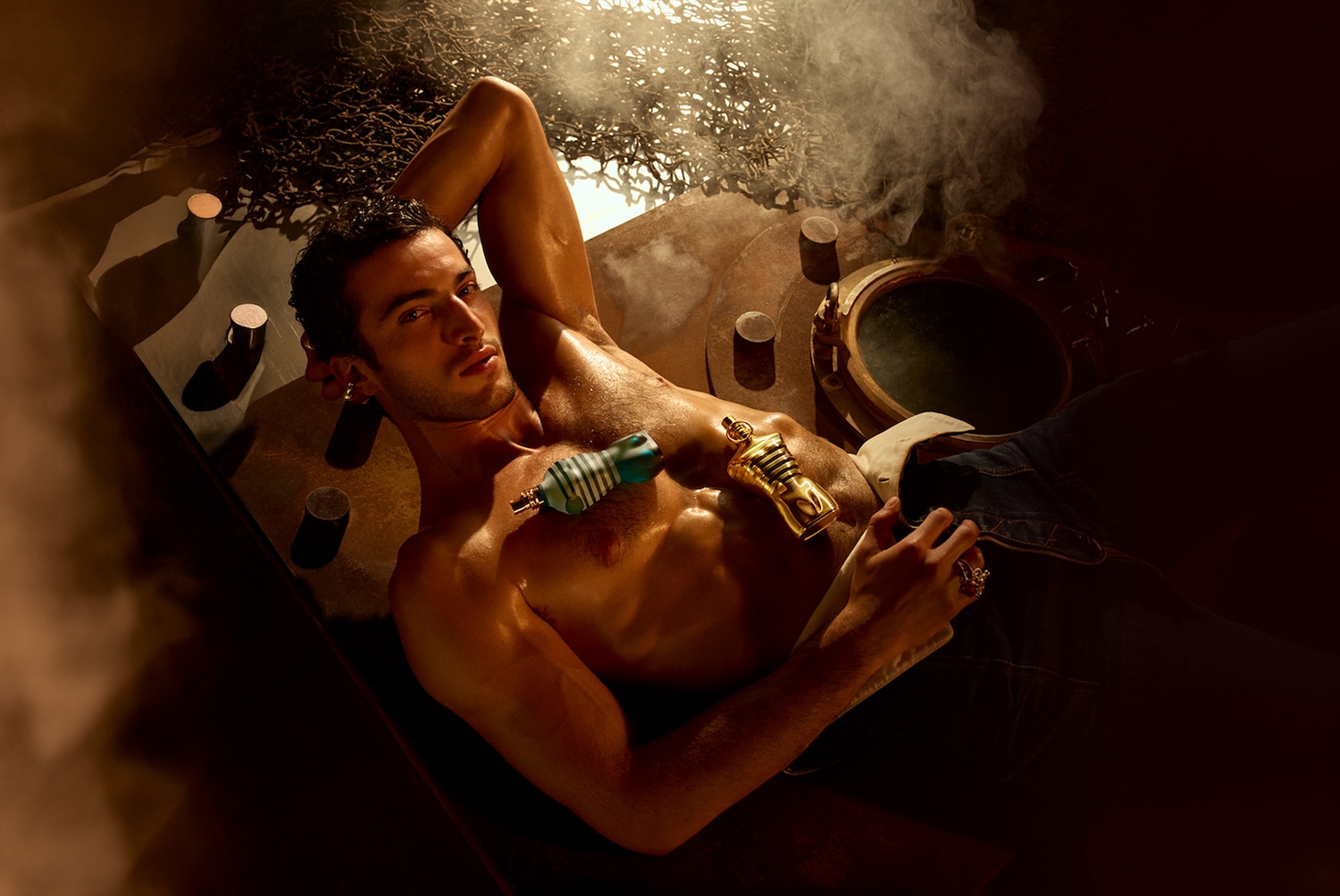 Ein Mann liegt mit freiem Oberkörper da, auf seiner Brust liegen zwei Flakons in Form eines Männerkörpers, eine goldene und eine blau-weiß gestreifte.