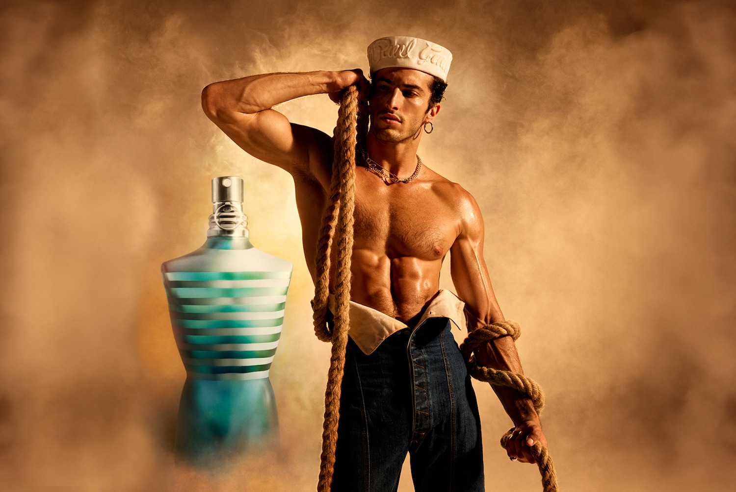 Ein muskulöser Mann mit freiem Oberkörper, Jeans und einer Kapitänsmütze posiert mit einem Seil neben einem Flakon in Form eines Männerkörpers mit blau-weissen Streifen.