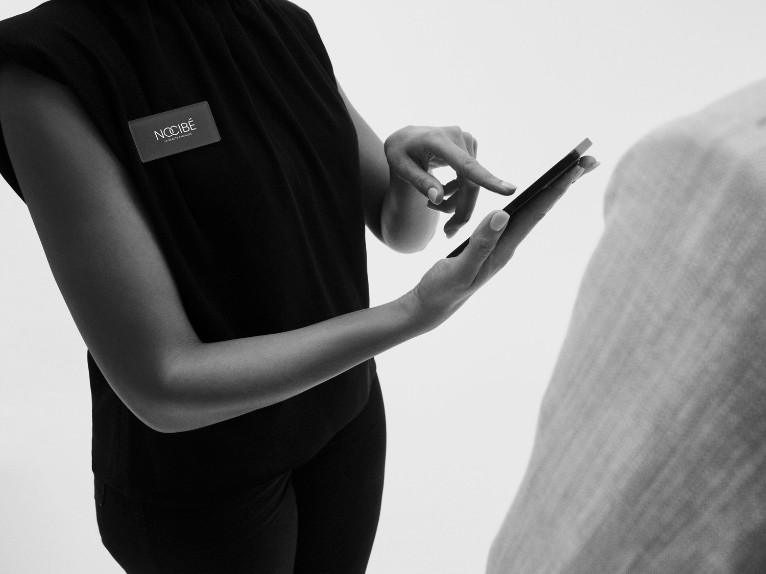 Une employée de Nocibé en uniforme noir utilise un smartphone, mettant en avant l'engagement de la marque envers la beauté et la technologie.