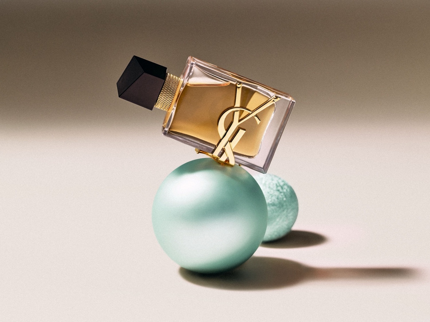 Ein YSL-Parfümflakon mit goldenem Inhalt und schwarzem Verschluss balanciert auf zwei blassgrünen Kugeln.