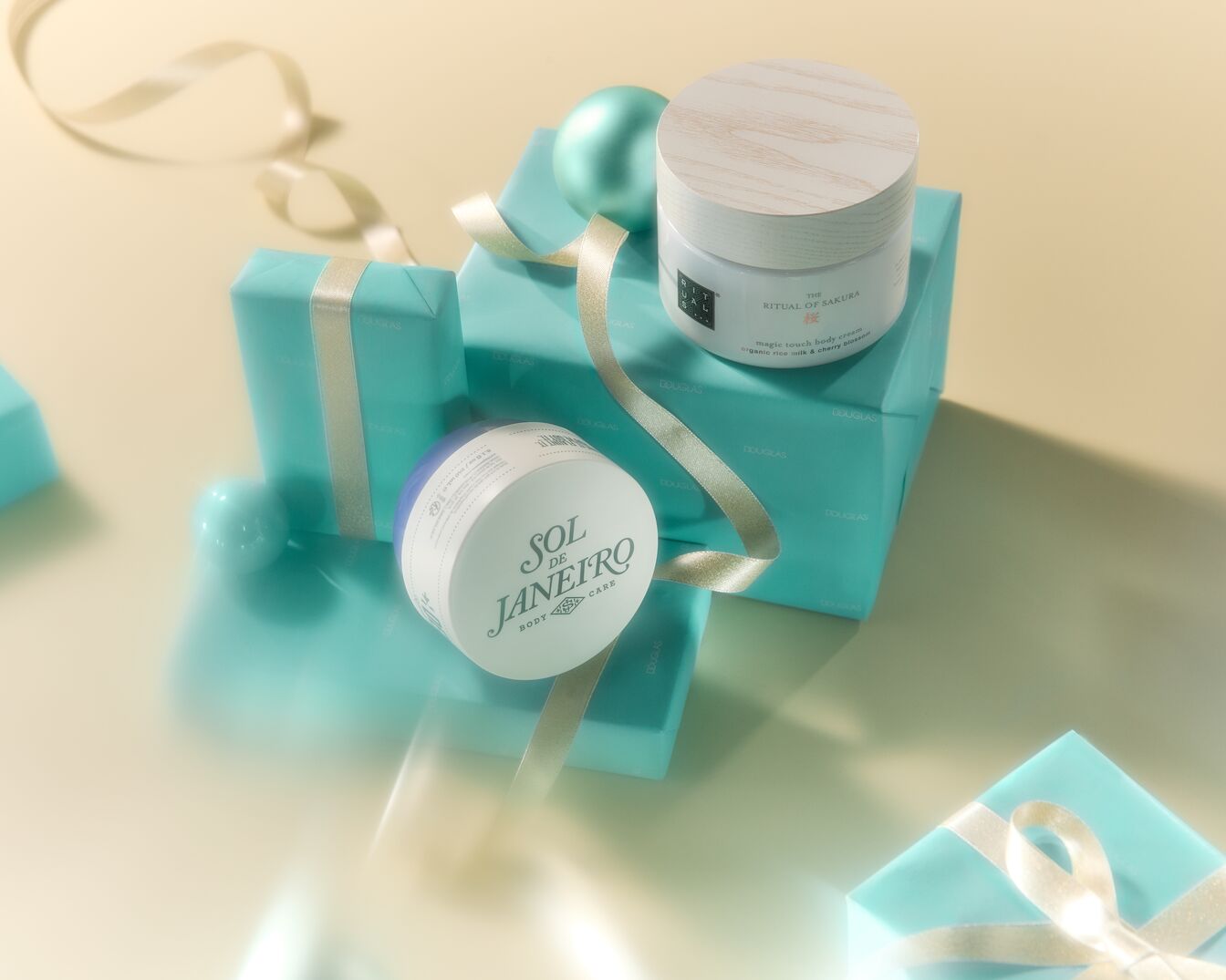 Šventinė kompozicija su „Sol de Janeiro“ kūno priežiūros priemone ir „Ritual of Sakura“ kūno kremu ant turkio spalvos dovanų dėžučių su auksiniais kaspinais.