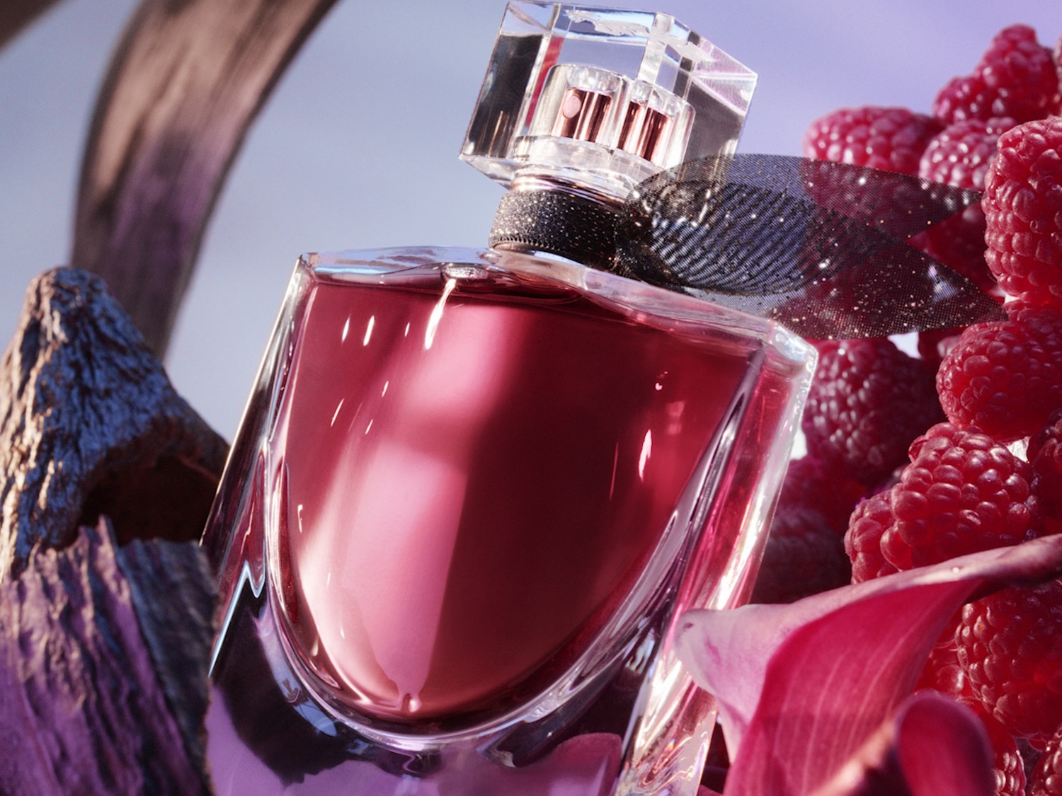 Stiklinis kvepalų buteliukas su juodu kaspinu, apsuptas aviečių, akmenų ir gėlės žiedo su rausvu skysčiu viduje.