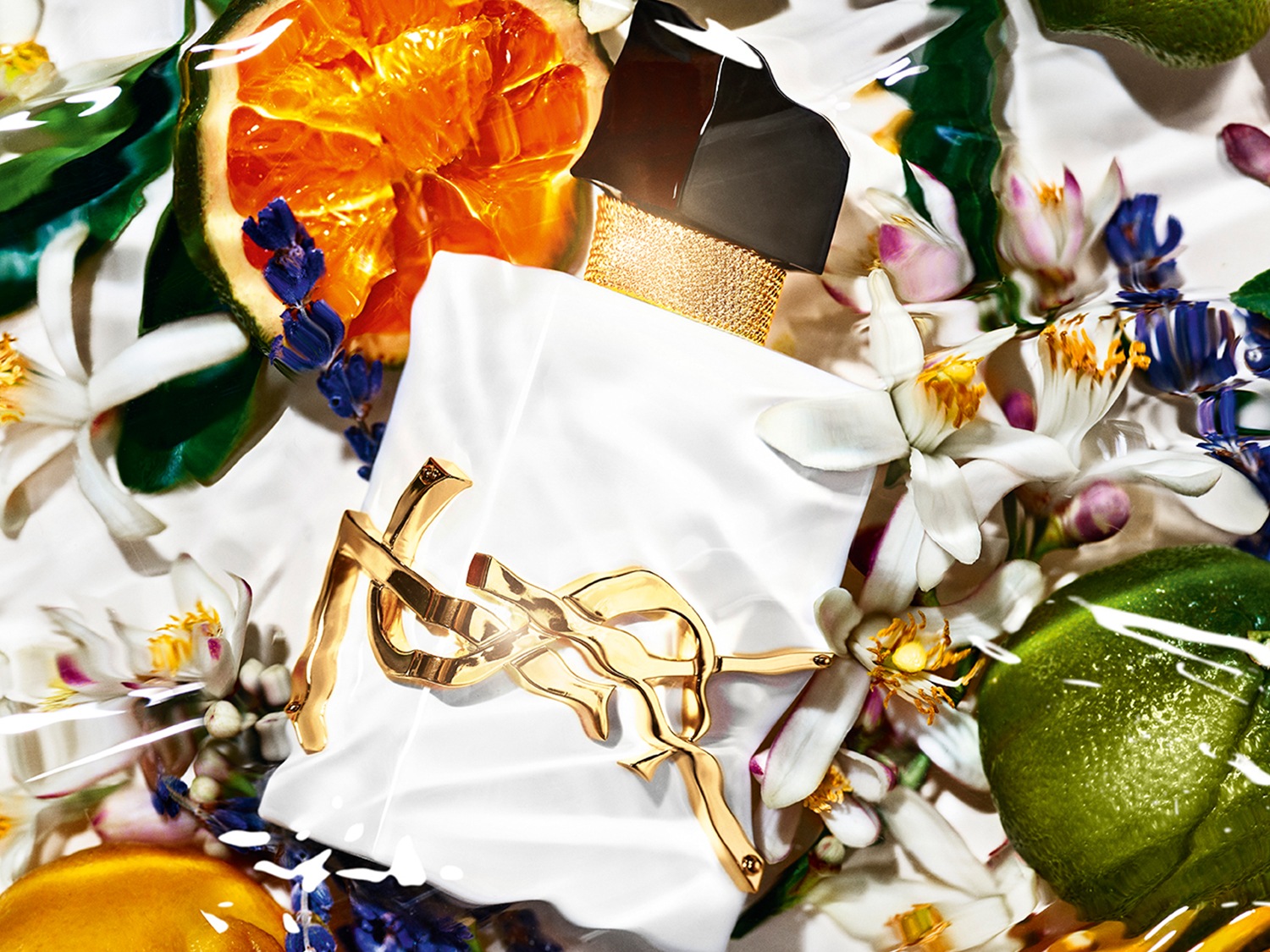 Baltas YSL kvepalų buteliukas su aukso spalvos logotipu, apsuptas apelsinų griežinėlių, žalių citrinų ir baltų žiedų, sukuria gaivų ir prabangų įvaizdį.