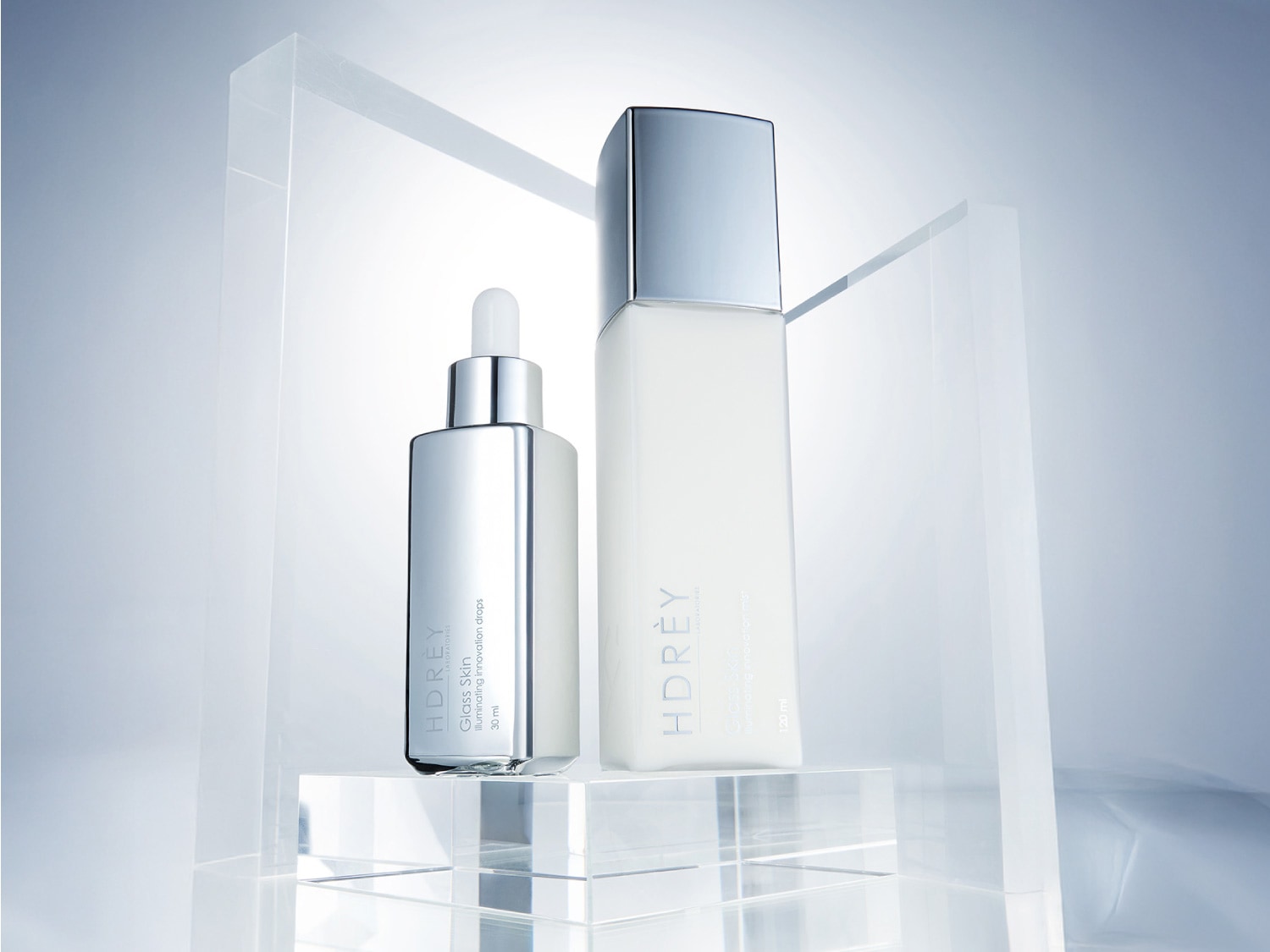 Dwa srebrne flakony HDREY Glass Skin: jeden z pipetą, drugi z dozownikiem, stojące na przezroczystym bloku, emanujące nowoczesnością i luksusem.