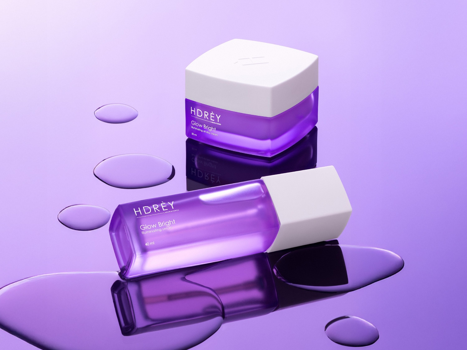 Fioletowy zestaw kosmetyków HDRÈY Glow Bright, zawierający rozświetlający krem na dzień w białym słoiczku i serum w przezroczystej butelce, oba z białymi nakrętkami, otoczone kroplami wody na fioletowym tle.
