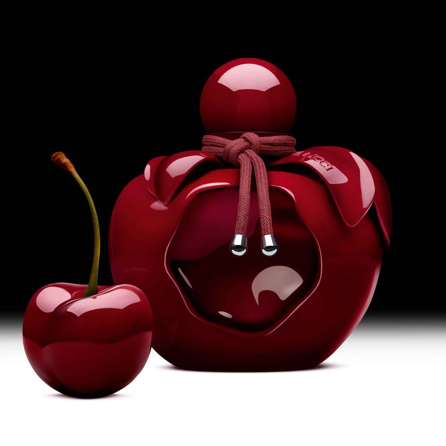 Un flacon de parfum rouge en forme de pomme est présenté à côté d'une cerise rouge brillante, évoquant un parfum fruité et gourmand.