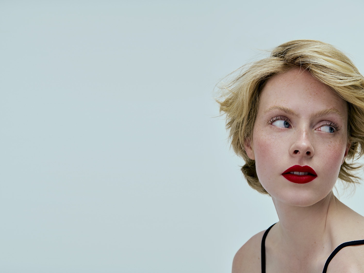 Primo piano di una donna con capelli biondi corti, lentiggini e labbra rosse che guarda di lato su uno sfondo chiaro.