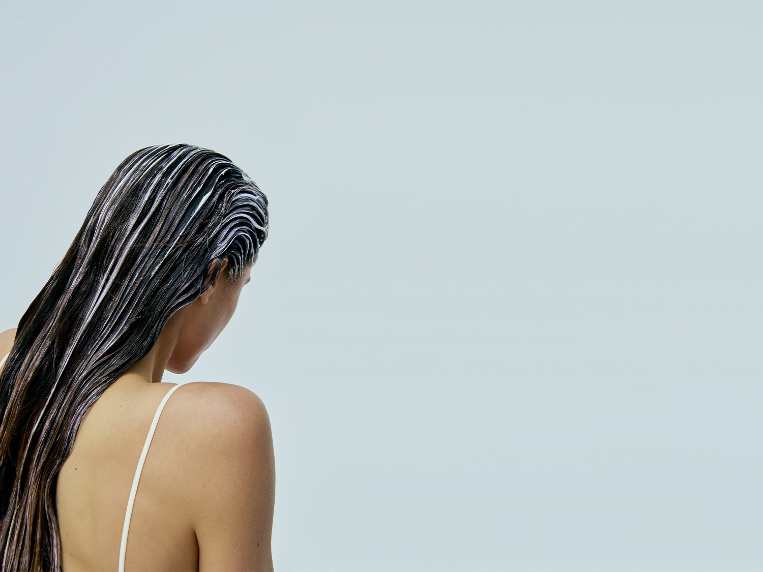 Vista da dietro di una donna con lunghi capelli castani, coperti da un trattamento bianco cremoso, che si prepara a nutrire e rivitalizzare la chioma.