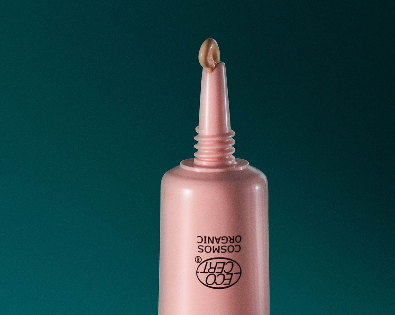 Nahaufnahme einer hellrosa Tube mit der Aufschrift 'COSMOS ORGANIC' und einem kleinen Klecks beiger Creme auf der Spitze, vor einem dunkelgrünen Hintergrund.