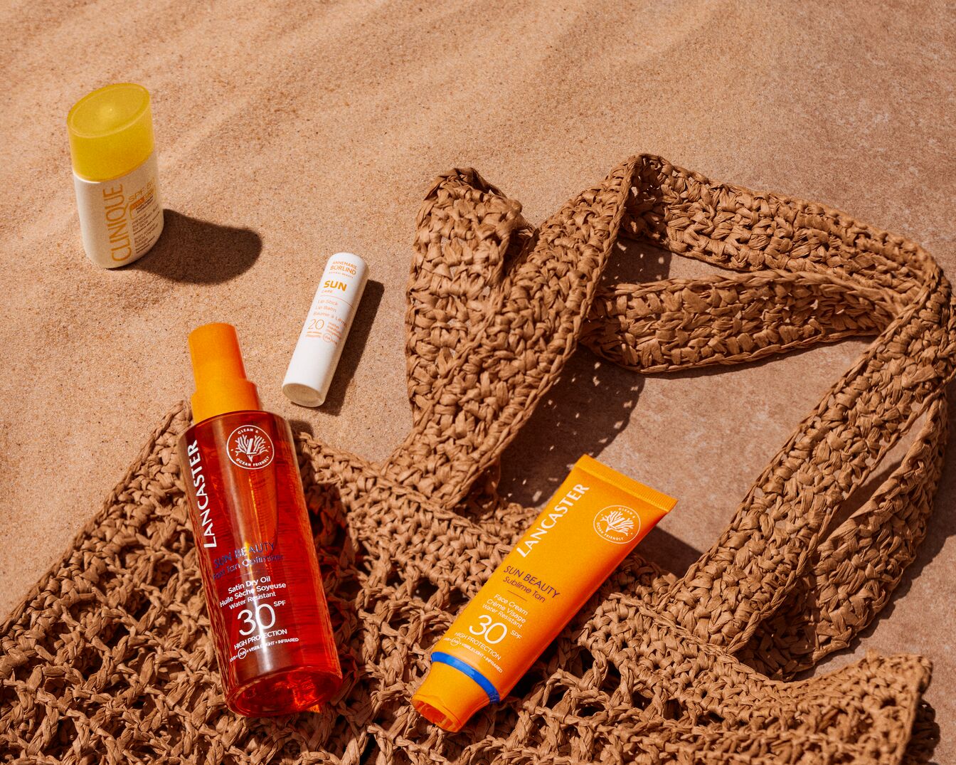 Sonnencreme von Clinique, Lancaster und Annemarie Börlind liegen auf einem beigen Strandtuch im Sand und bieten hohen Schutz mit LSF 30 und 20.