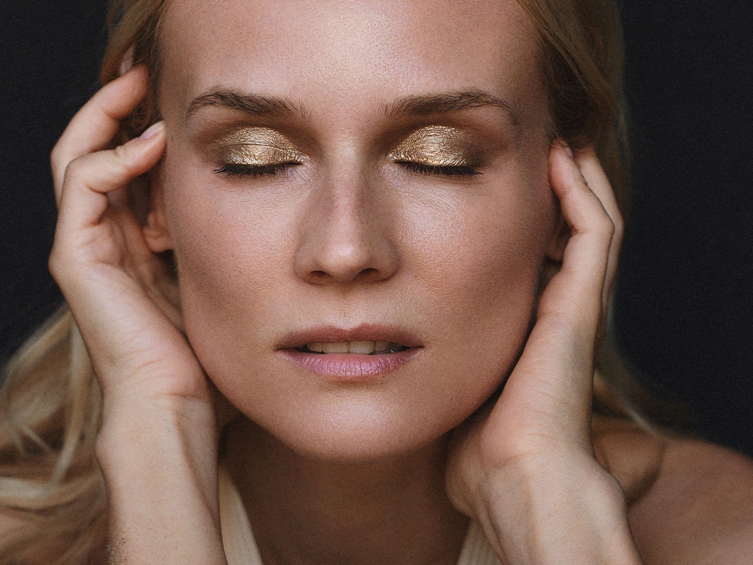 Šviesiaplaukė moteris užmerktomis akimis, ant vokų tepa auksiniai akių šešėliai, rankos prigludusios prie veido.