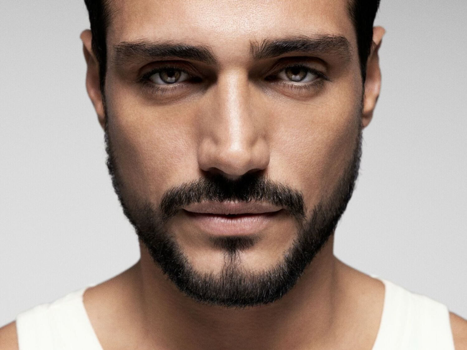 Artimas jauno vyro portretas su tamsiais plaukais, rudomis akimis ir barzda, žiūrintis tiesiai į priekį.