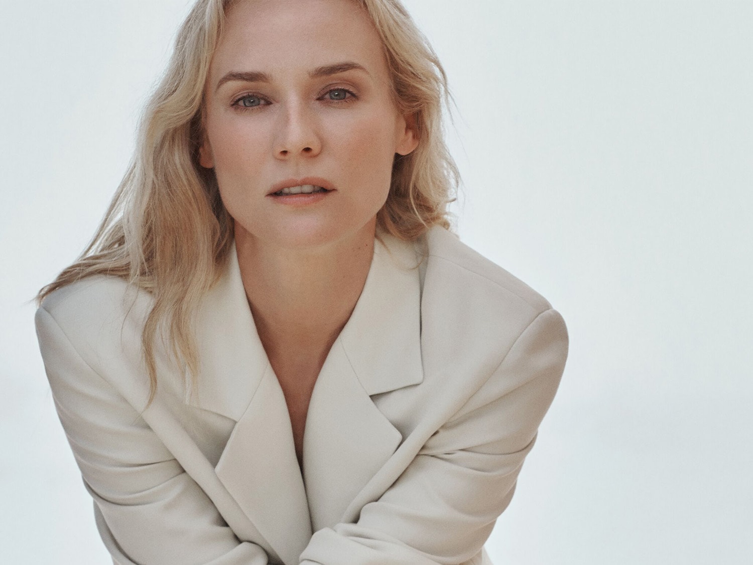 Šviesiaplaukė moteris su šviesiu švarku, žiūrinti tiesiai į priekį, sukuria ramybės ir elegancijos įspūdį.