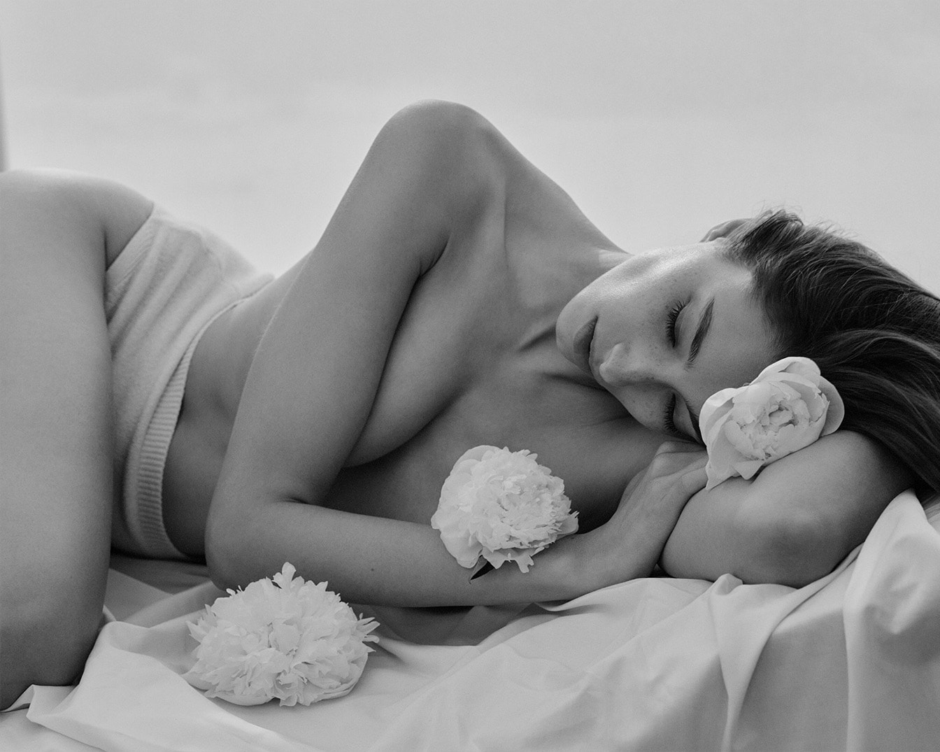 En noir et blanc, une femme se repose, les yeux clos, entourée de pivoines blanches, évoquant une sérénité naturelle et une beauté pure.