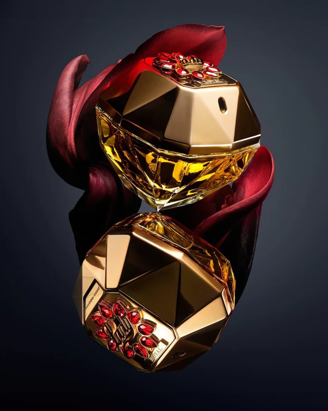 Nahaufnahme von zwei goldenen Parfümflaschen in Diamantenform, die mit roten Edelsteinen verziert sind und auf einer reflektierenden Oberfläche mit roten Blütenblättern liegen.