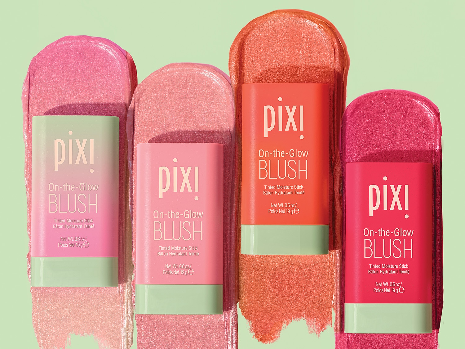 Cztery sztyfty Pixi On-the-Glow BLUSH w odcieniach różu i pomarańczy, każdy z zieloną podstawą i napisem z nazwą produktu, leżą obok siebie na zielonym tle.