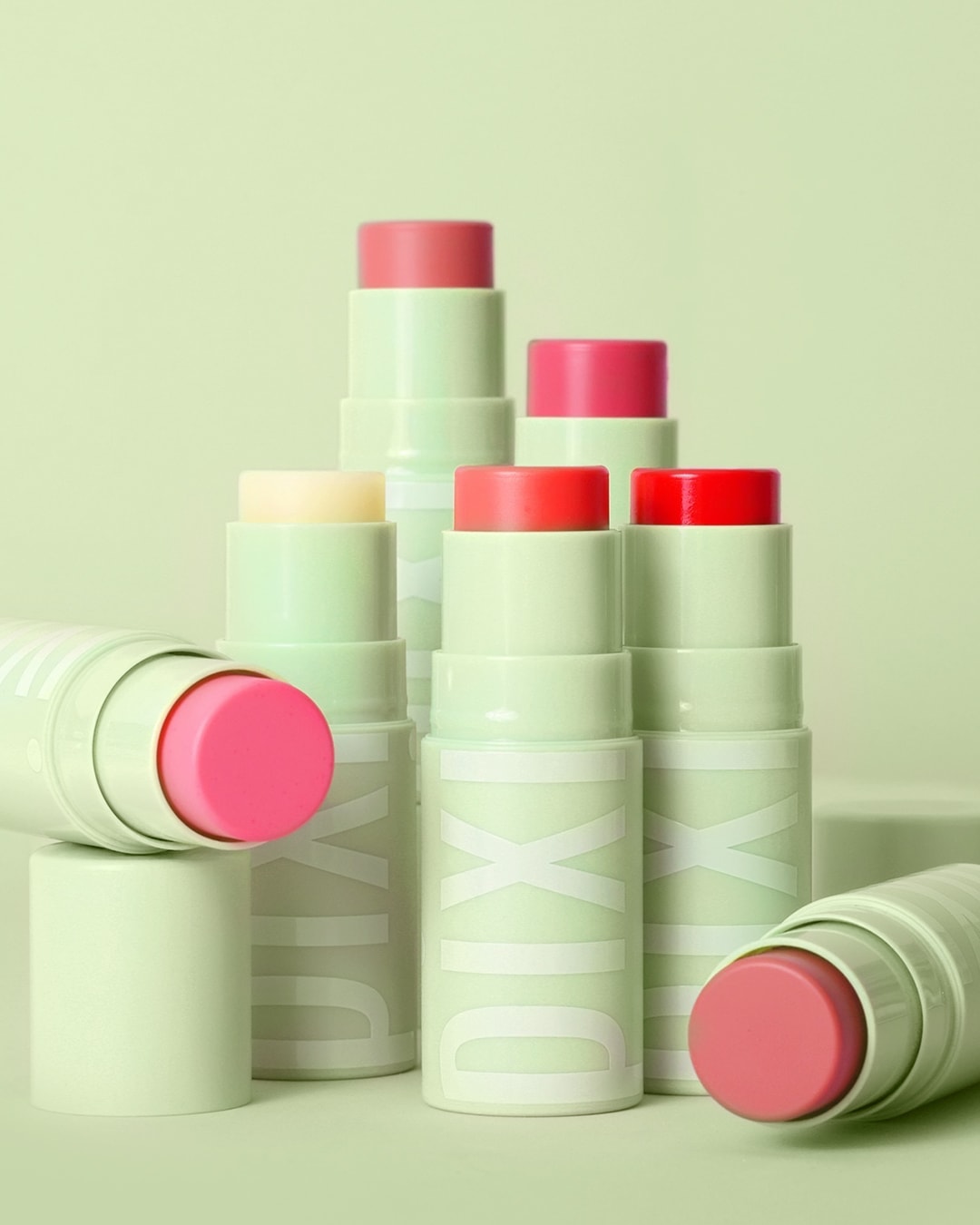 Na jasnozielonym tle ustawione są balsamy do ust Pixi w sztyfcie, każdy w innym odcieniu różu i czerwieni, tworząc barwną kompozycję.