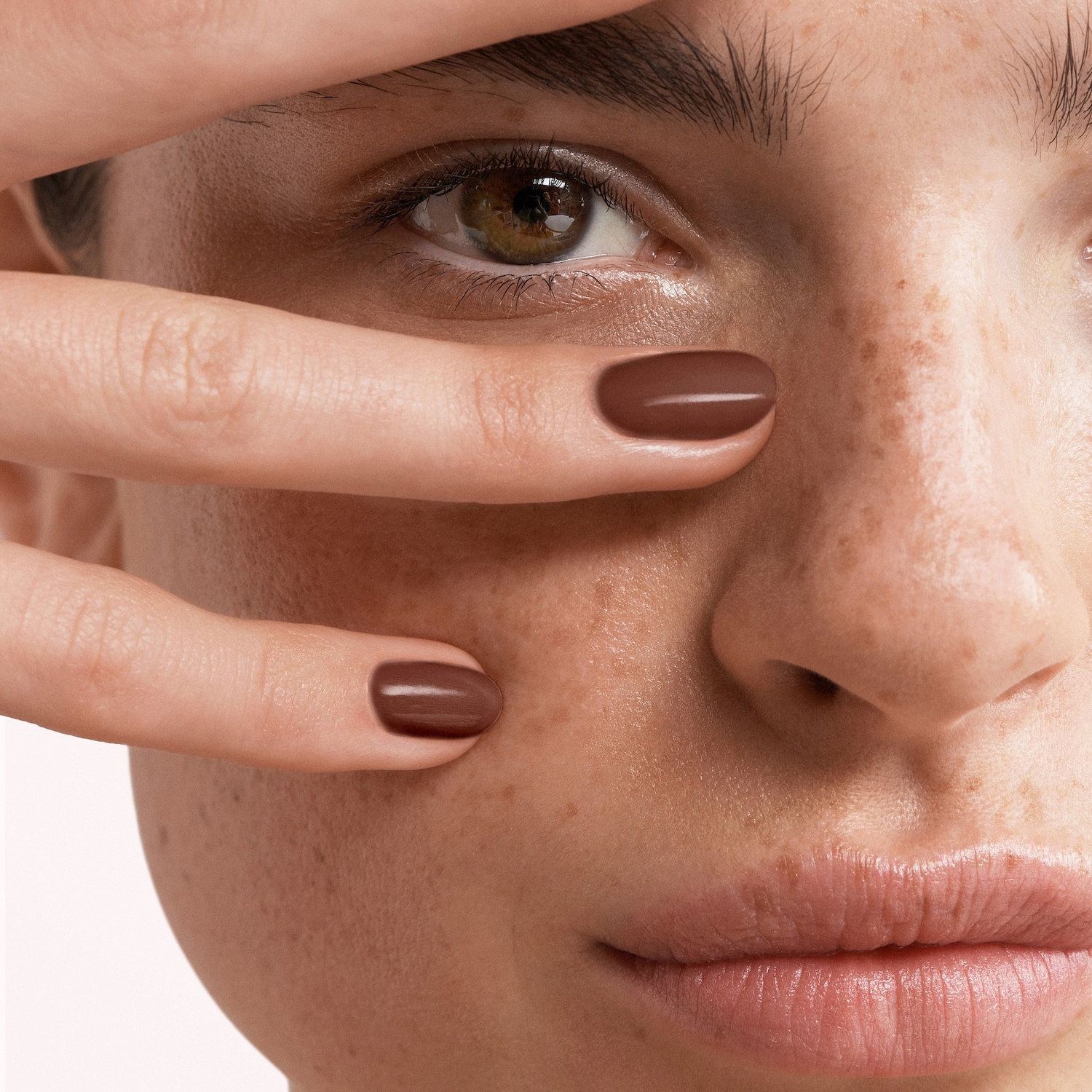 Gros plan sur le visage d'une femme avec des taches de rousseur, ses doigts ornés d'un vernis à ongles marron chocolat encadrant son œil vert.