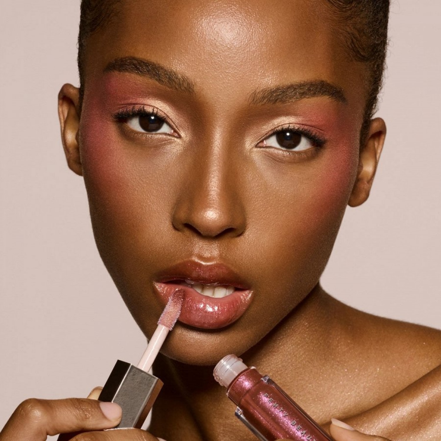 Een vrouw met een glanzende roze lipgloss van Fenty Beauty, die de glans aanbrengt met een applicator, terwijl haar make-up bestaat uit roze blush en oogschaduw.