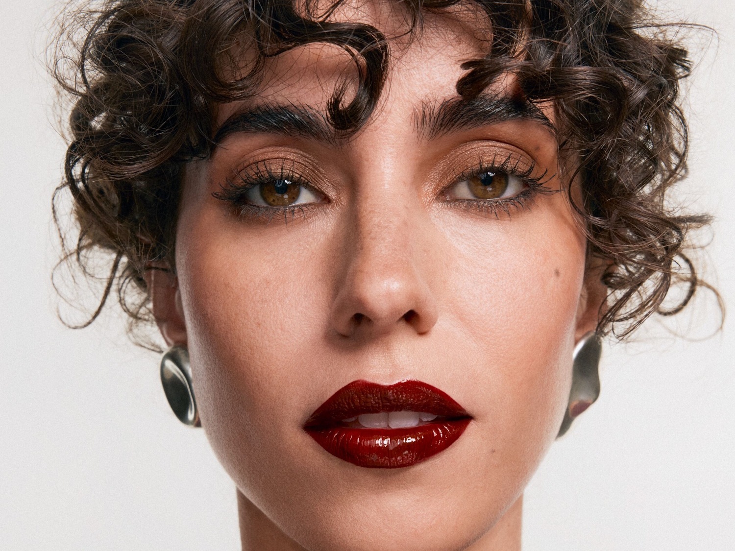 Een close-up van een vrouw met krullend bruin haar, glanzende bordeauxrode lippenstift en glinsterende oogschaduw, die haar gezicht en schouders vult.