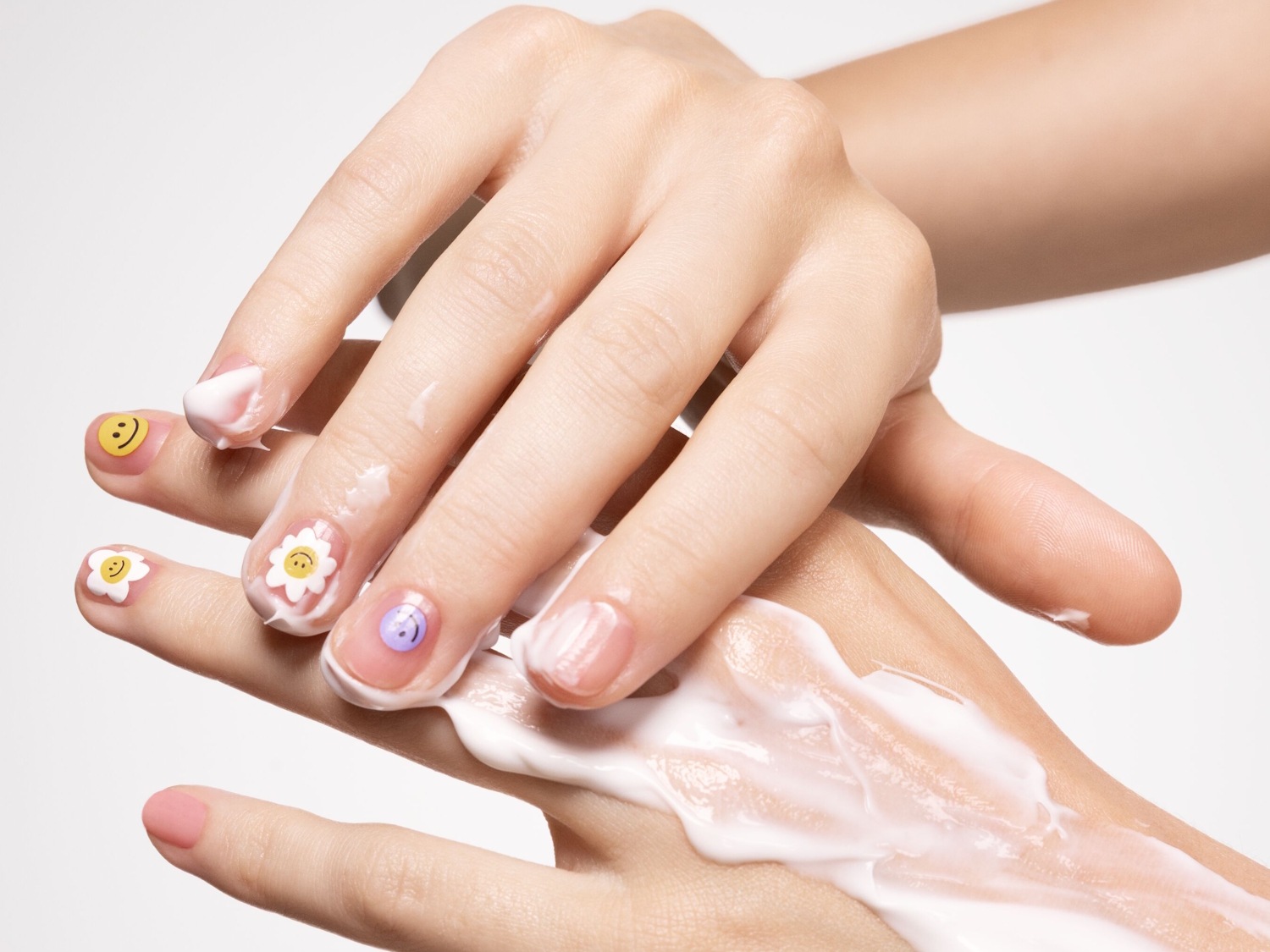 Nahaufnahme einer Hand, die mit einer anderen Hand eine weiße Creme verreibt, wobei die Nägel mit verschiedenen Aufklebern wie Smileys und Blumen verziert sind.
